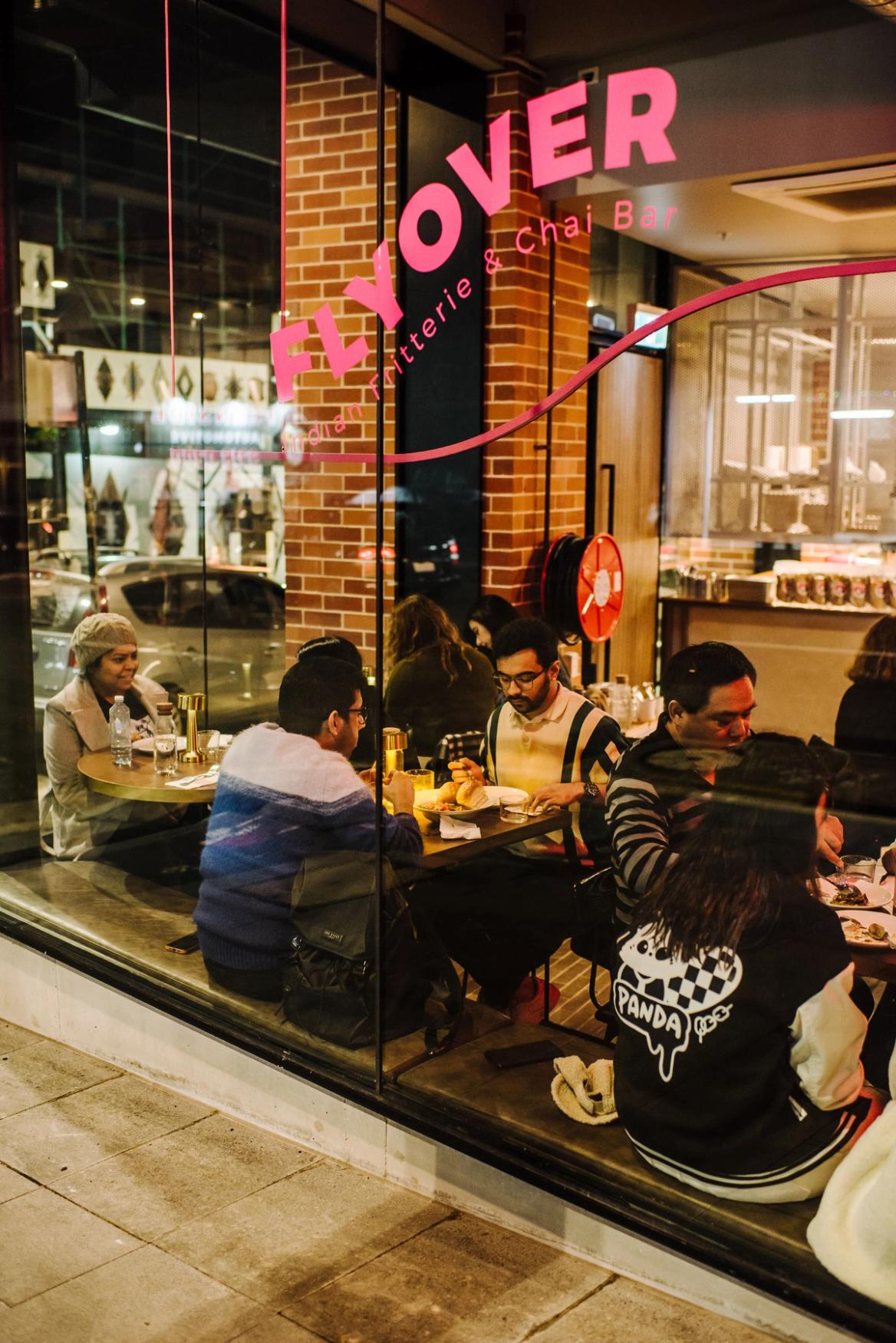 A cozy scene of people dining inside "Flyover Indian Fritterie & Chai Bar," viewed from the outside through a large glass window.