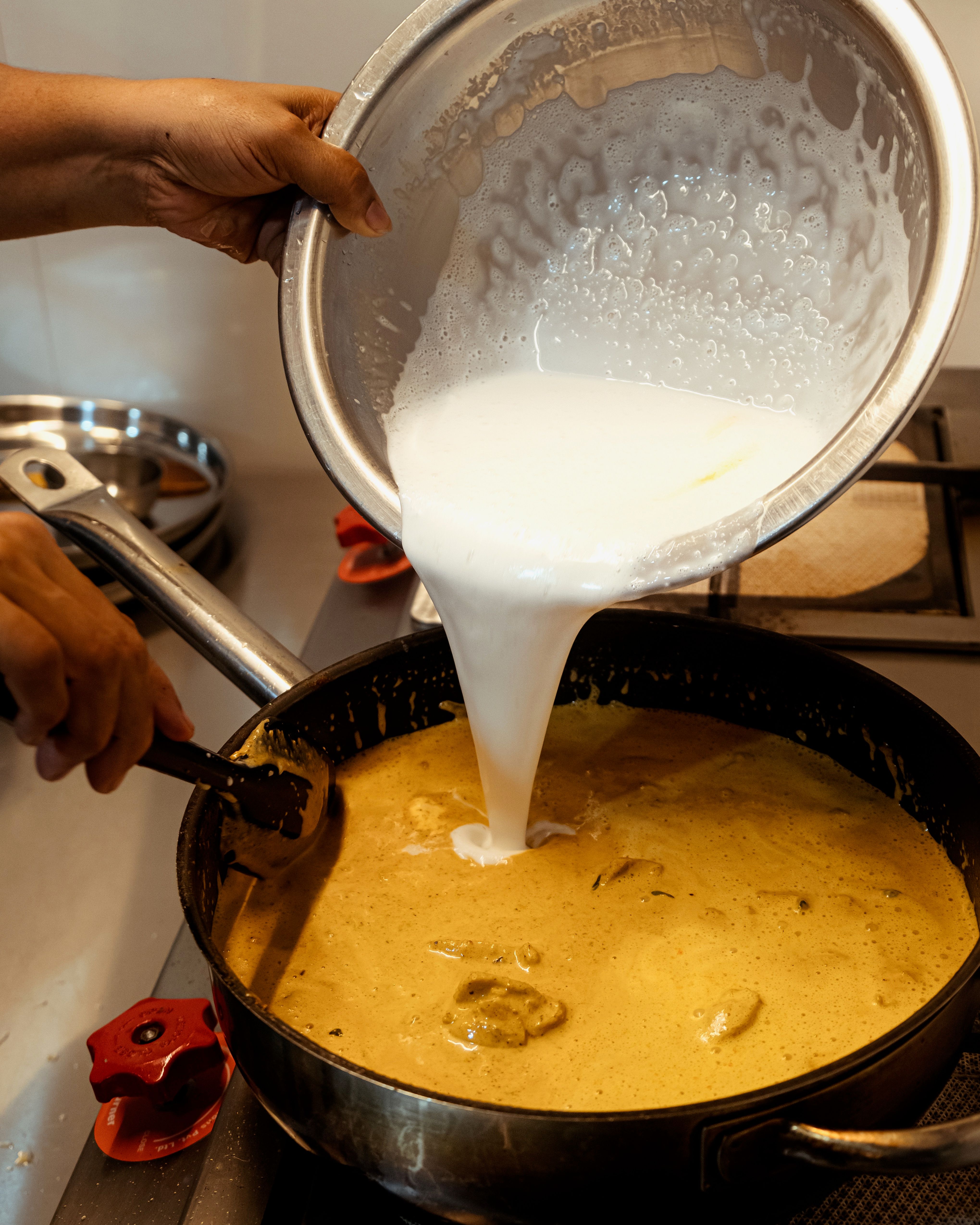 Brown female hands pour coconut milk from a large stainless-steel bowl into a stove-top pan with chicken and curry ingredients.