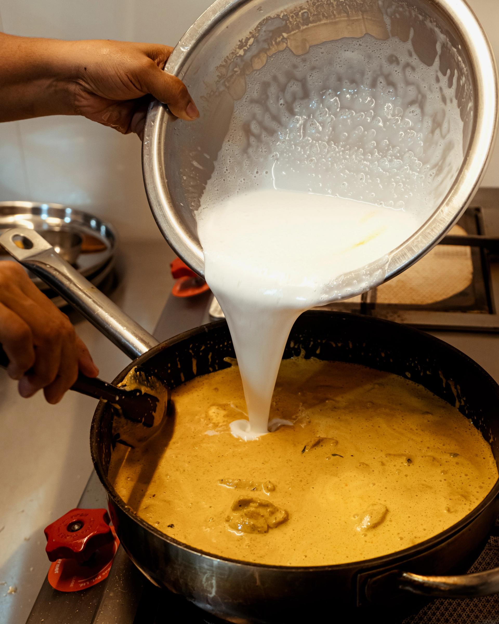 Brown female hands pour coconut milk from a large stainless-steel bowl into a stove-top pan with chicken and curry ingredients.
