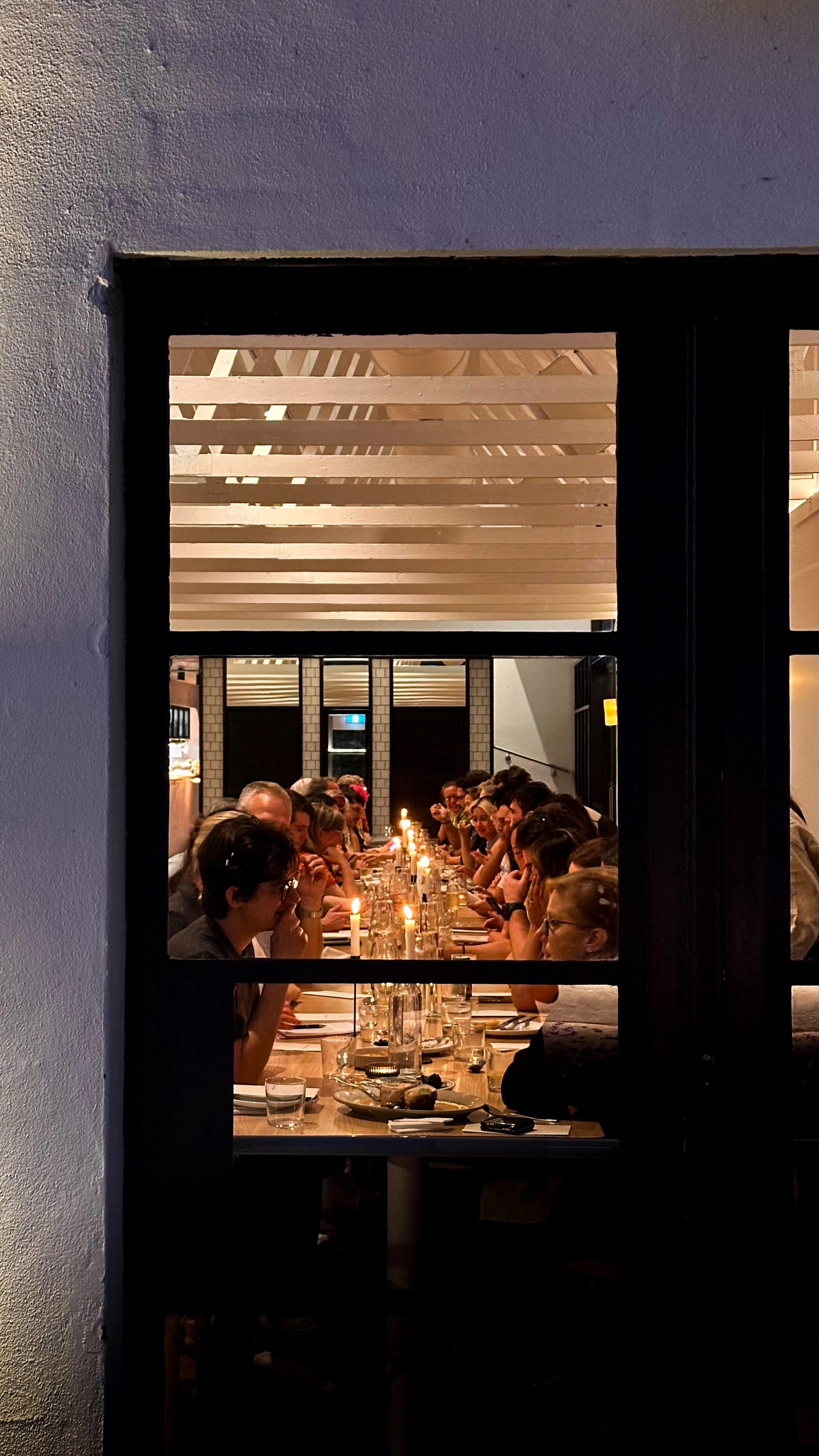 A long table of people talking over a candle lit dinner, viewed from outside the window.