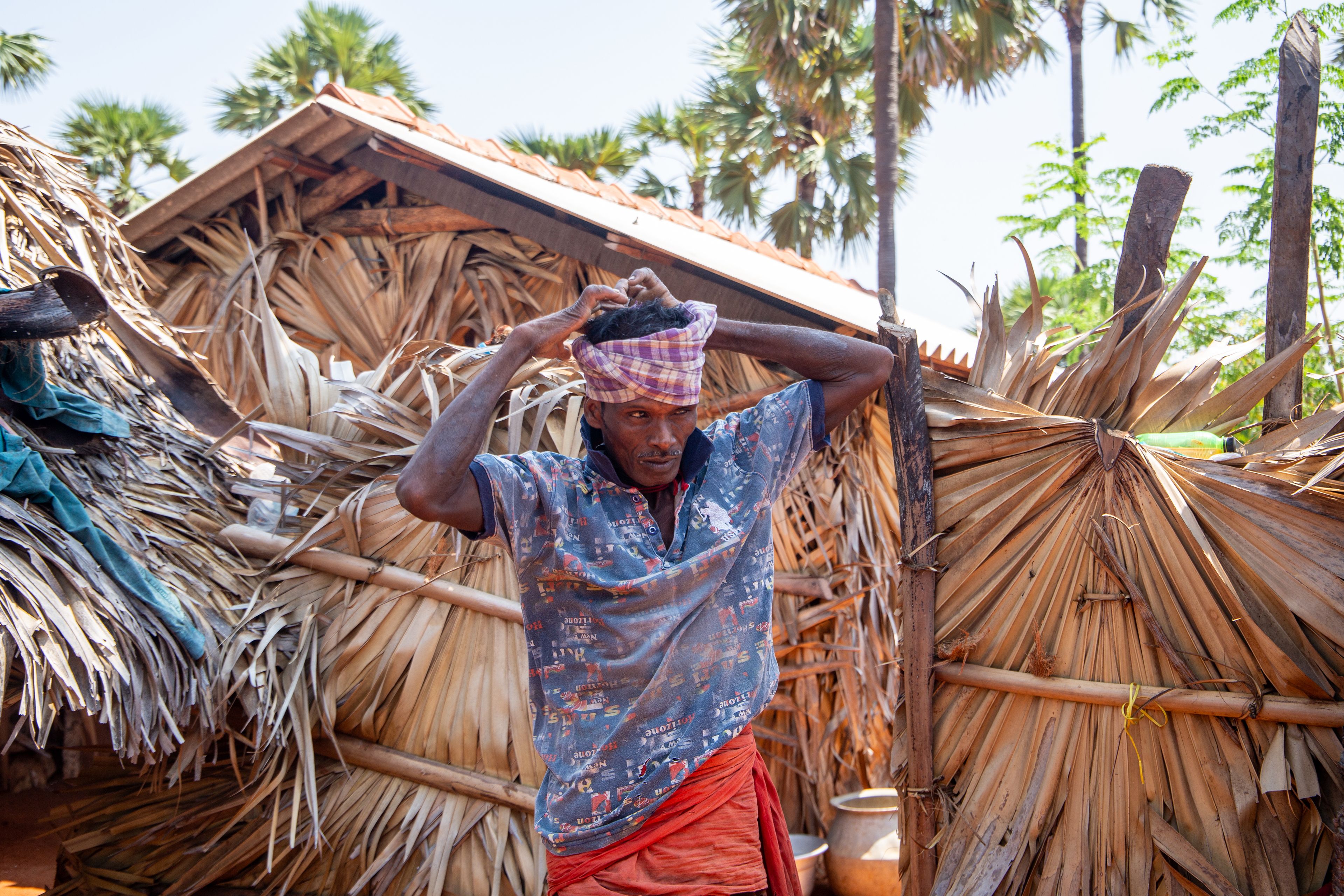A Tamil palm tree climber stands in front of a wall of dried palm leaves. He ties a red bandana around his forehead.