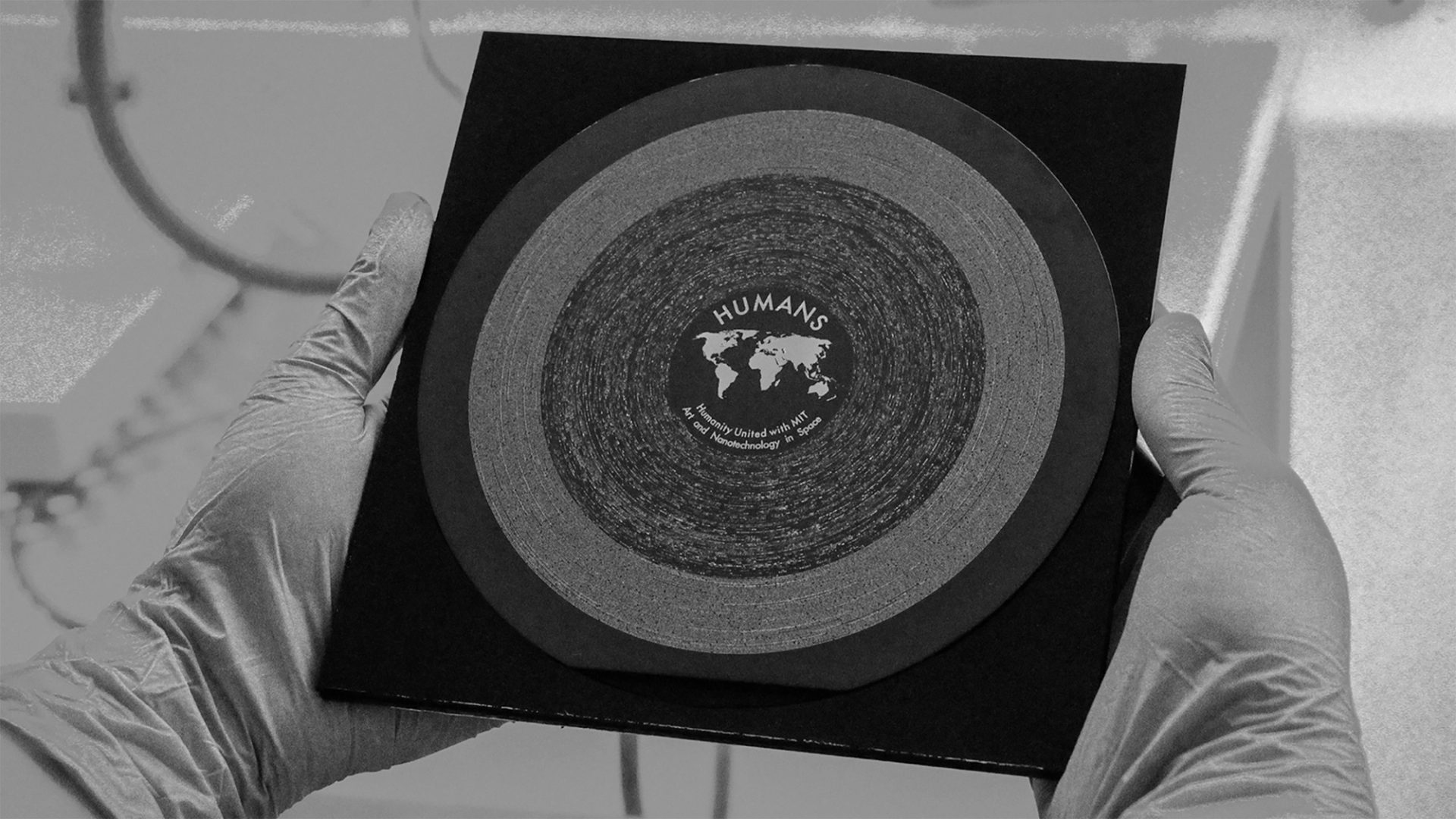 Black and white image of gloved hands holding a square sleeve carrying the HUMANS vinyl, a circular disc made with nanotechnology and white etchings.