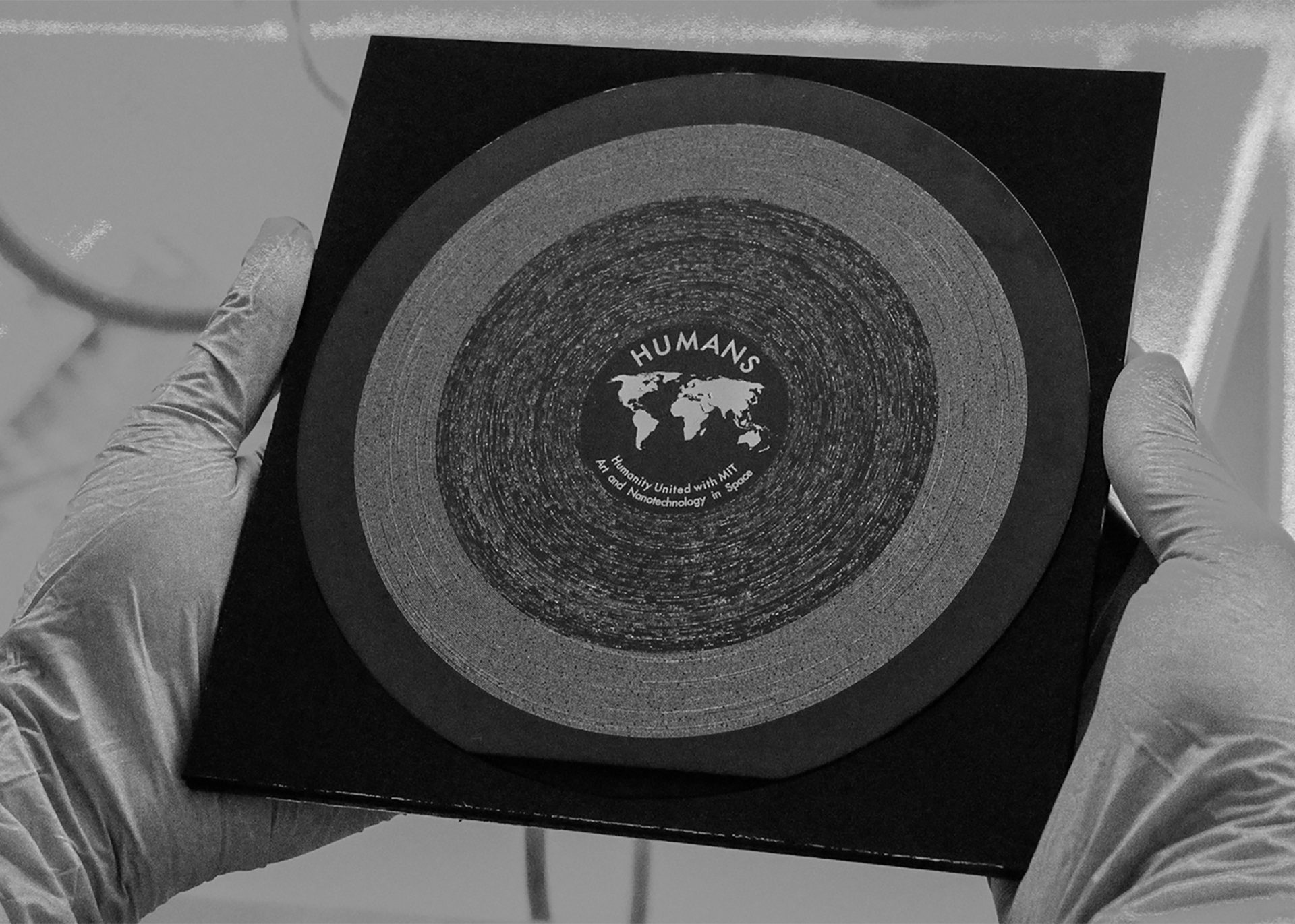 Black and white image of gloved hands holding a square sleeve carrying the HUMANS vinyl, a circular disc made with nanotechnology and white etchings.