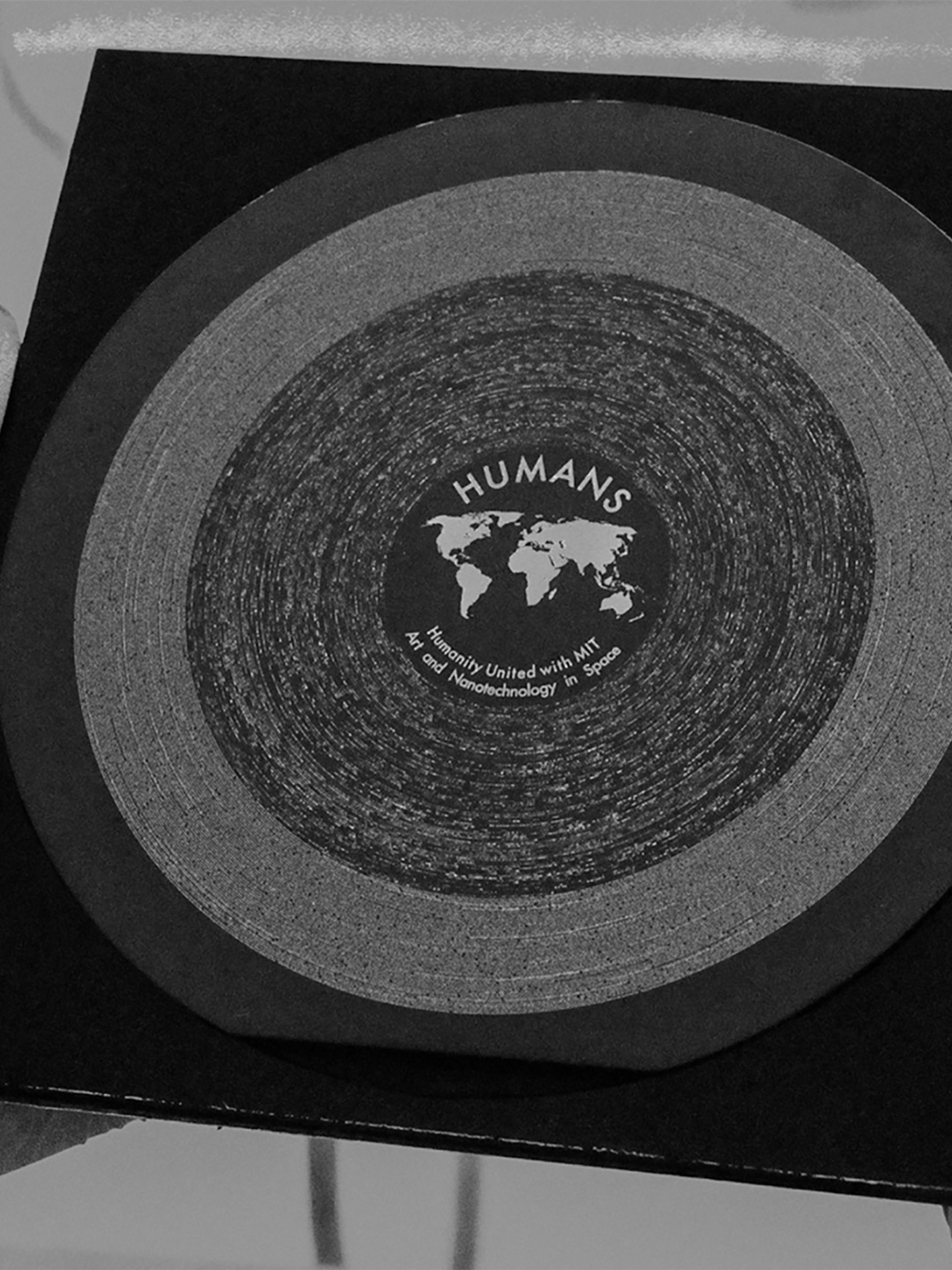 Black and white image of gloved hands holding a square sleeve carrying the HUMANS vinyl, a circular disc made with nanotechnology and white etchings.