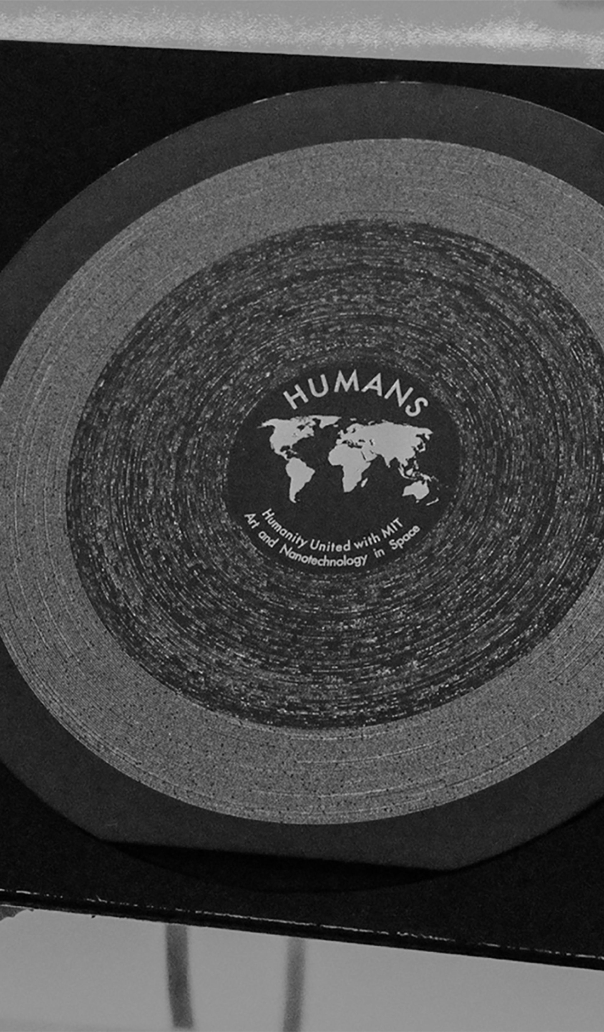Black and white image of gloved hands holding a square sleeve carrying the HUMANS vinyl, a circular disc made with nanotechnology and white etchings.