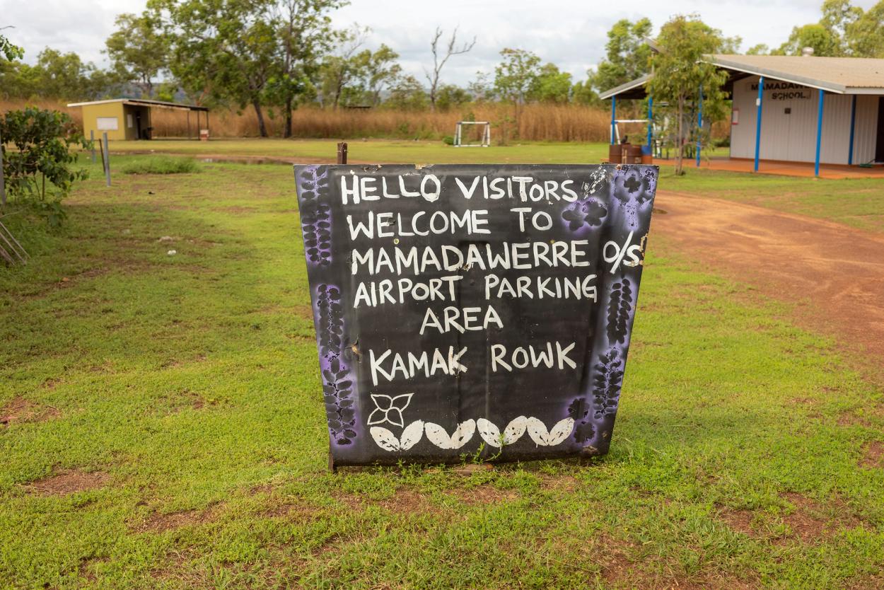 A black sign with purple and white illustrations bordering the text, ‘HELLO VISITORS, WELCOME TO MAMADAWERRE O/S AIRPORT PARKING AREA KAMAK ROWK’