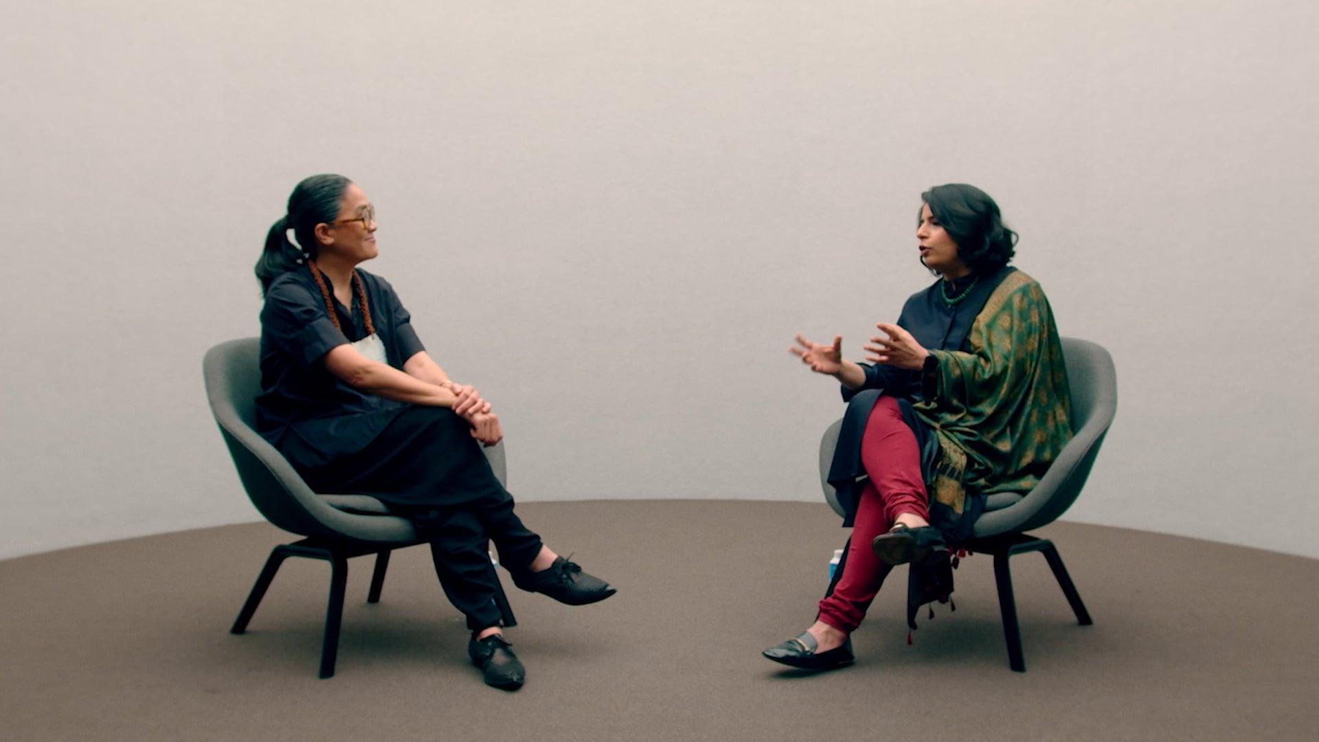 Two figures sitting on chairs in conversation.