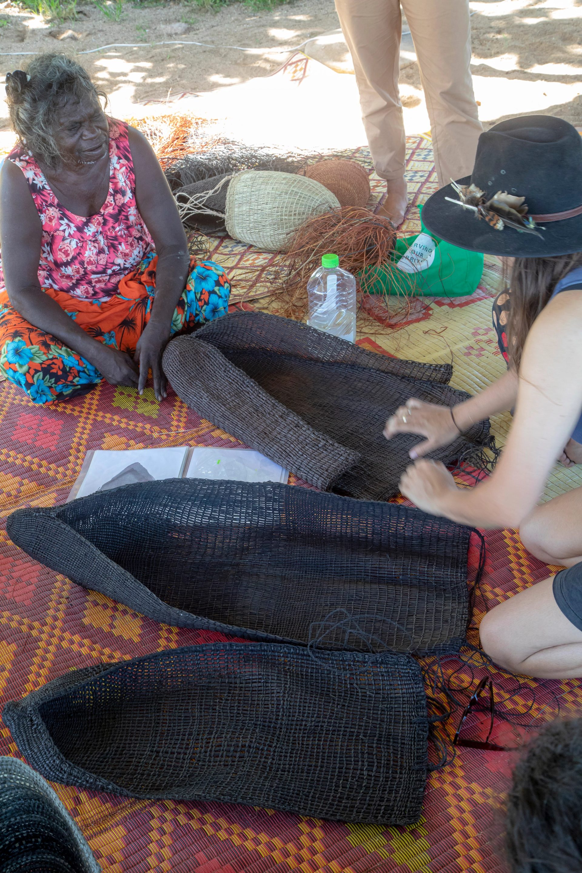 Older and younger woman sit talking across woven mats and baskets