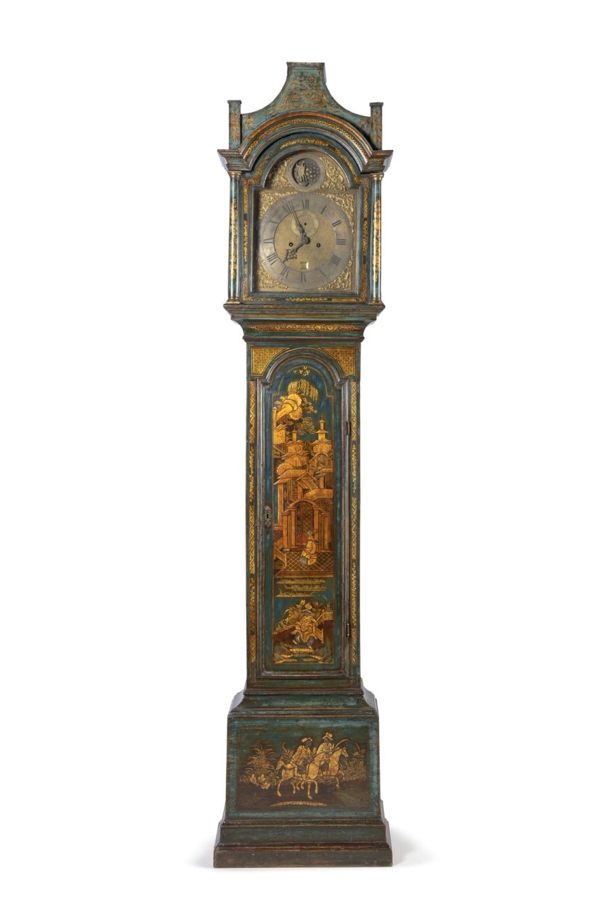 Ornate metal clock on a tall wood and metal stand carved with images of a village and people riding horses.