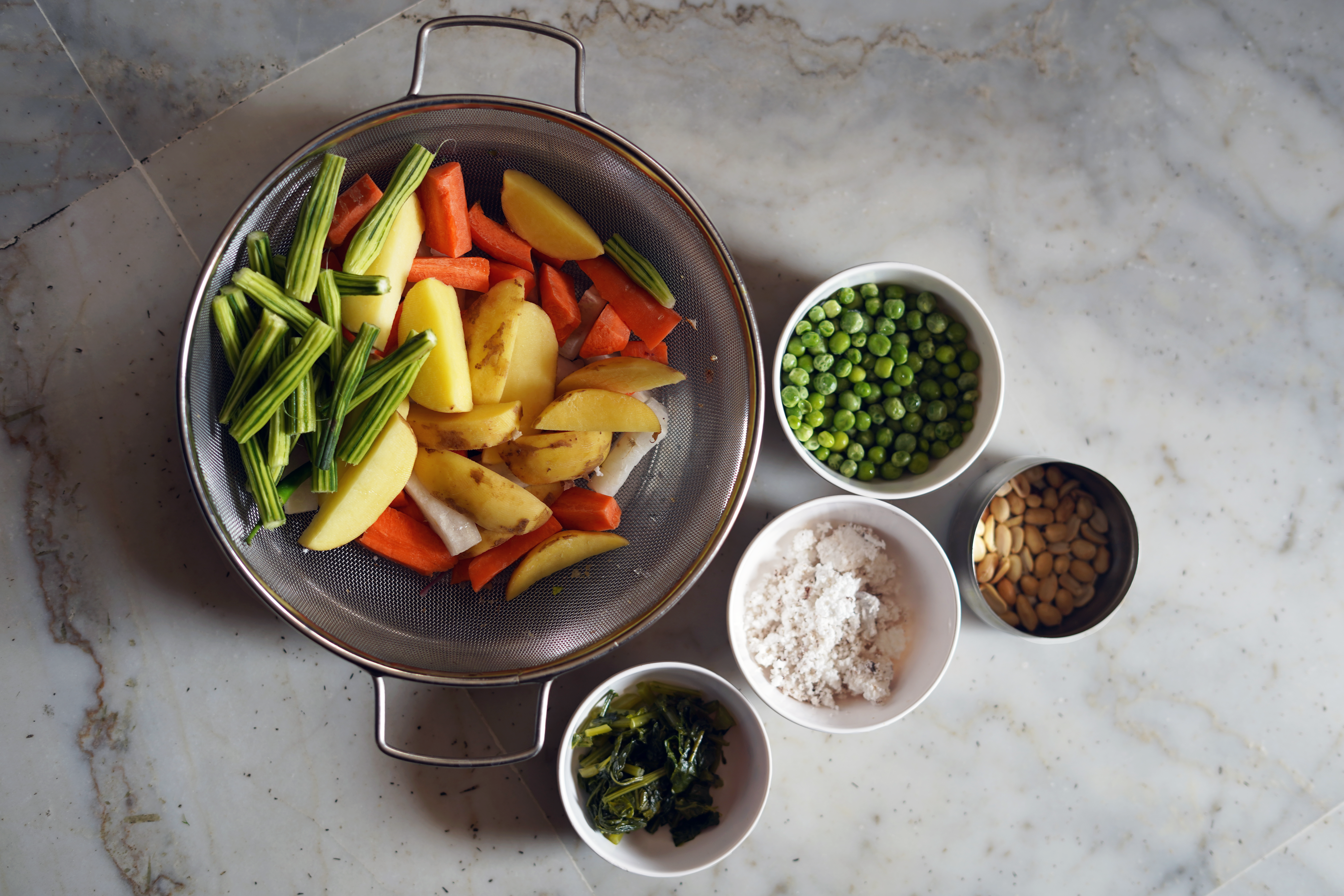 A large silver pot filled with green moringa pods, potato and carrot slices; four smaller bowls with peas, coconut, peanuts and radish leaves on a white marble-pattern surface.