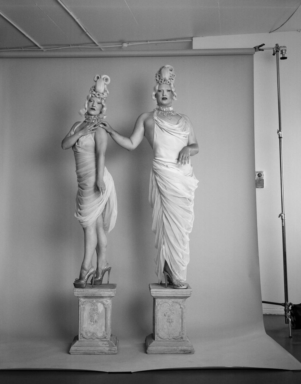 A black and white photograph of two people dressed in white draped dresses standing on pillars in a photography studio.