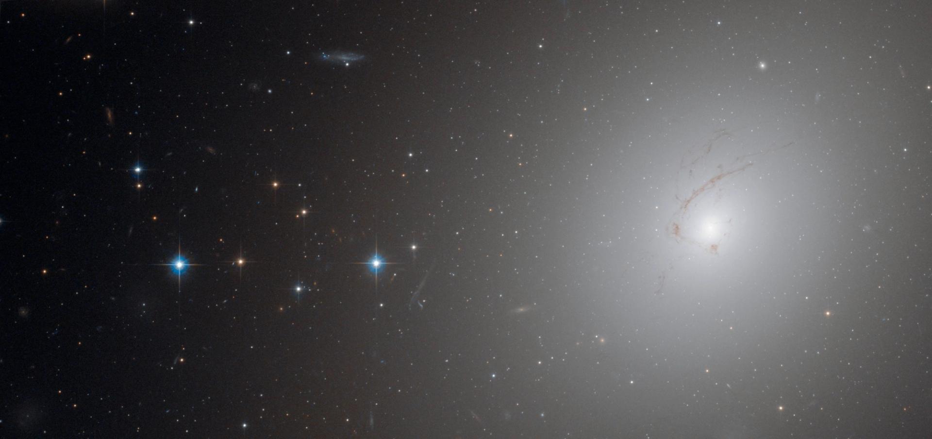 Photograph taken by Hubble’s Advanced Camera, shows NGC 4696, the largest galaxy in the Centaurus Cluster. With a bright white light coming from the right, and cluster of stars on the left.