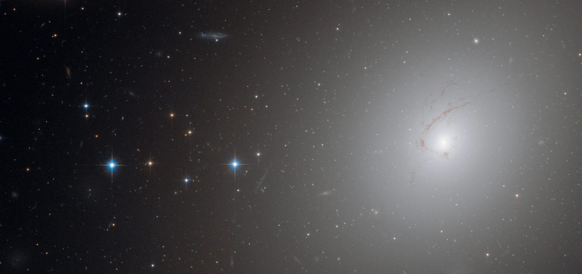 Photograph taken by Hubble’s Advanced Camera, shows NGC 4696, the largest galaxy in the Centaurus Cluster. With a bright white light coming from the right, and cluster of stars on the left.