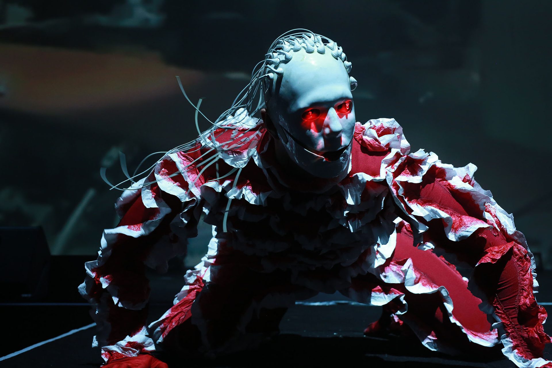 Justin Shoulder is Carrion - a creature in a white mask with red eyes and headphone cords as strands of hair. Carrion wears a red and white ruffled outfit. He is in motion.