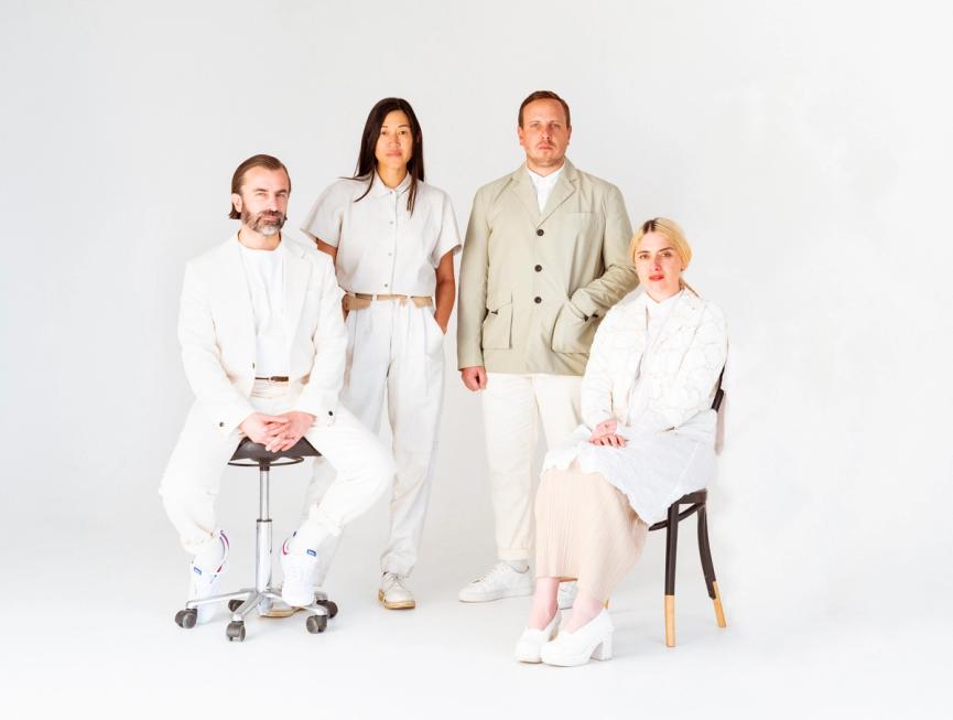 Four people dressed in white clothing sit and stand in front of a white backdrop.