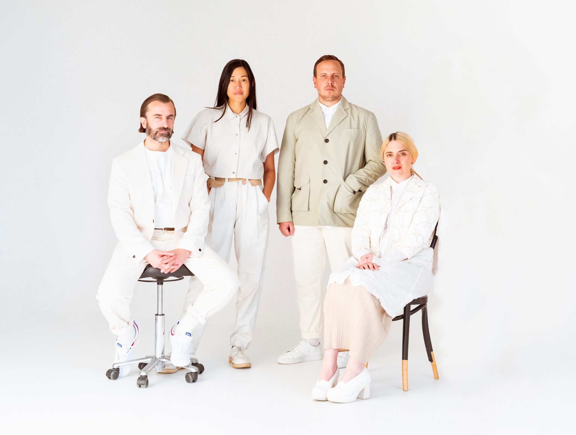 Four people dressed in white clothing sit and stand in front of a white backdrop.