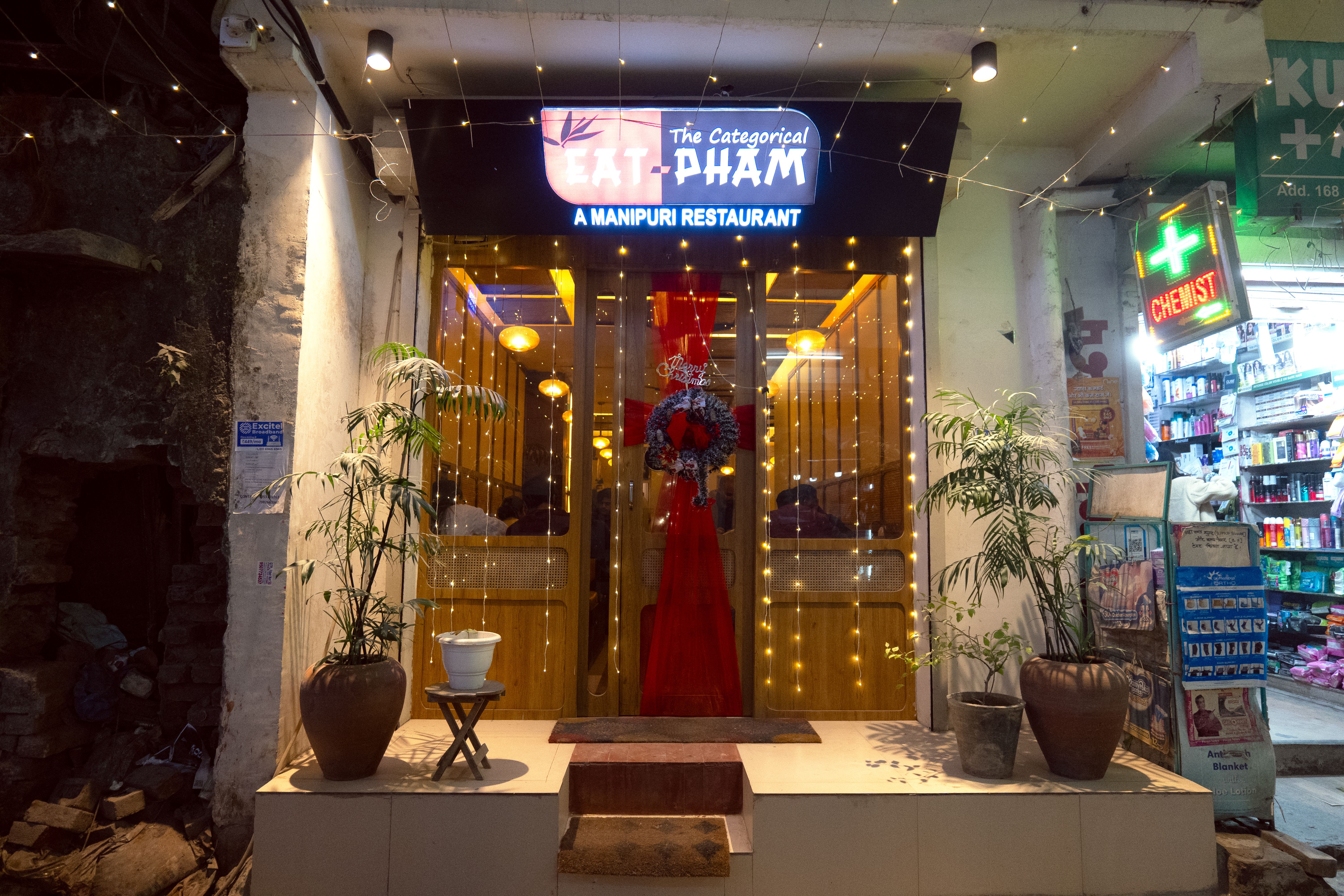 The exterior of a restaurant with a half red and half black lightbox sign reading ‘EAT PHAM’ and below, ‘A MANIPUR RESTAURANT’. Two plants are on either side of the entrance. Hanging lights are in the front windows and red fabric is styled across the door.