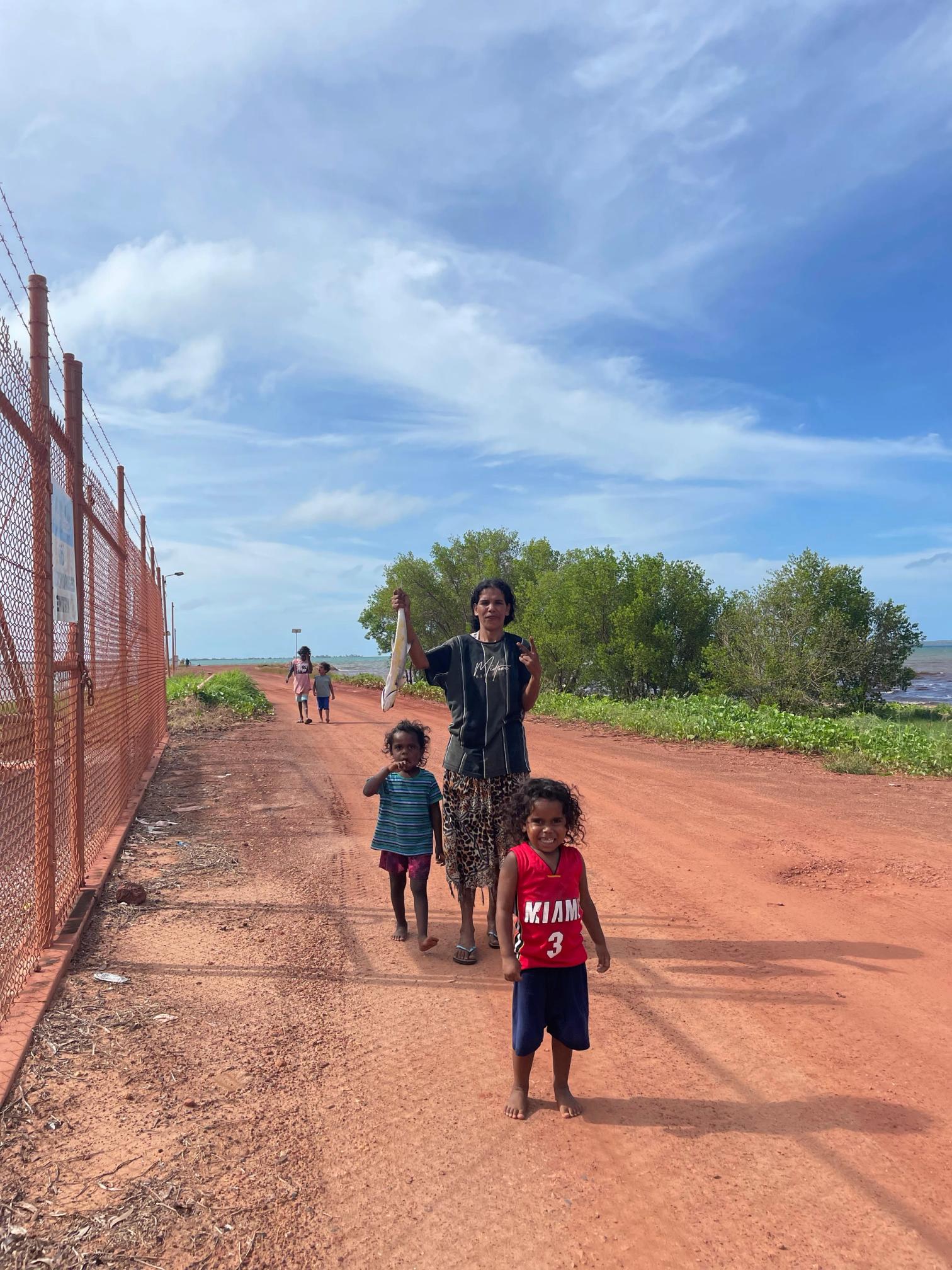A woman holds a freshly caught fish in her right arm. Next to her and in front of her are two small children. On the dirt orange road behind her are two children following along. The sky is blue and the ocean is visible far in the background.