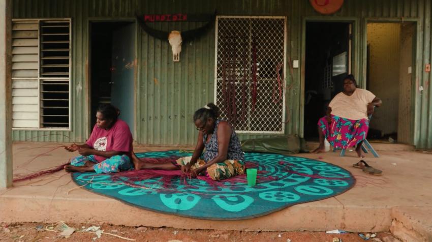 Women touching a traditionally woven mat on atop fabric on the floor.