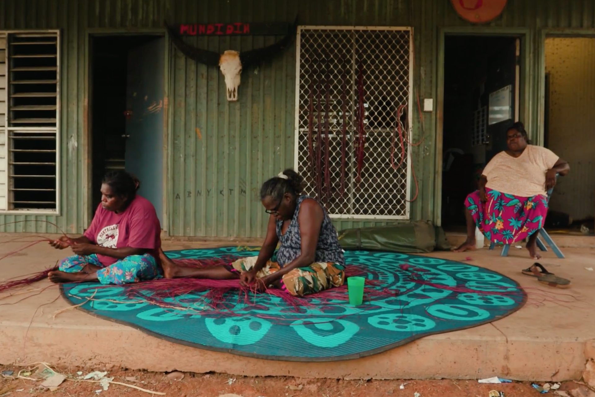 Women touching a traditionally woven mat on atop fabric on the floor.