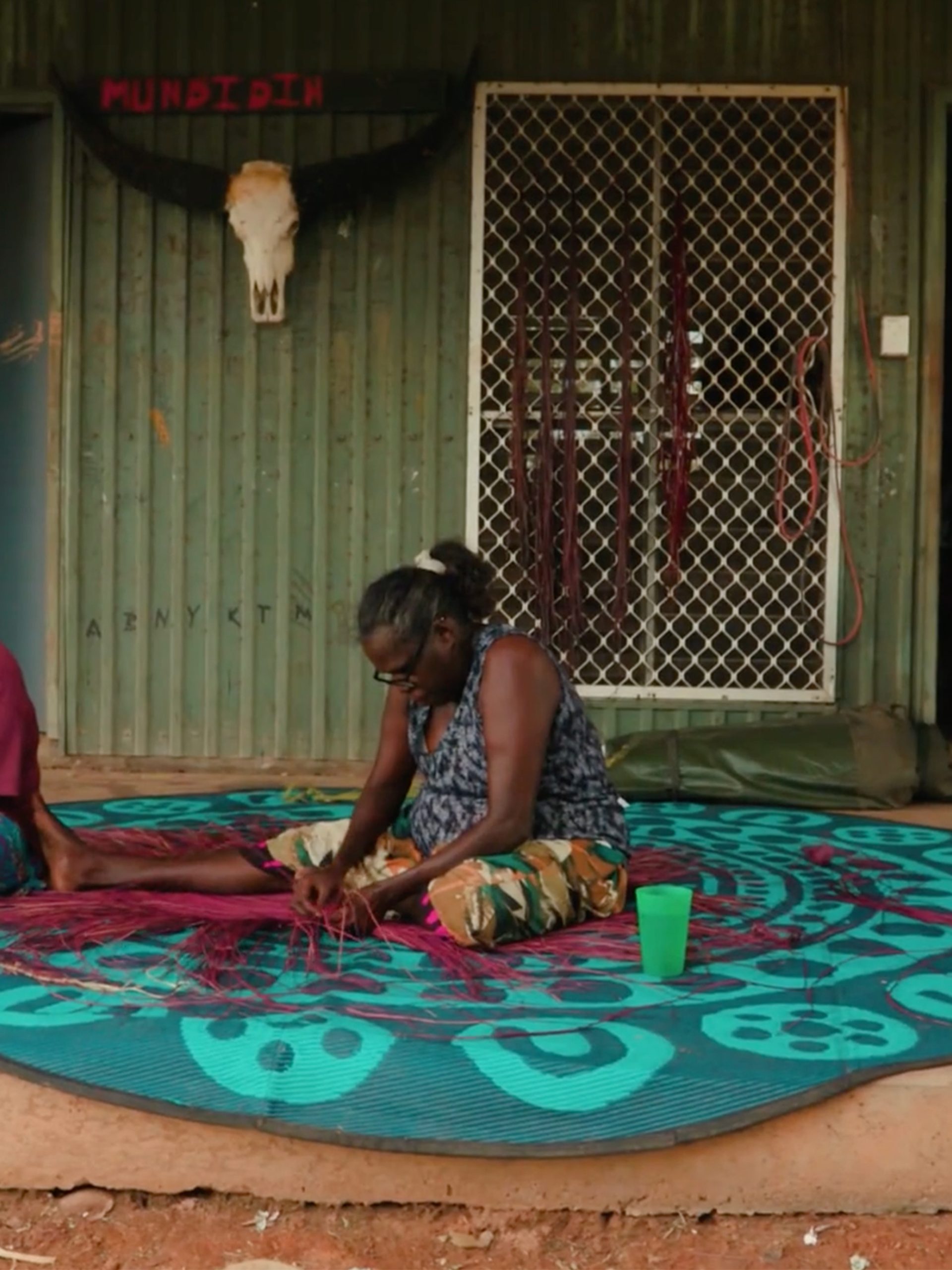 Women touching a traditionally woven mat on atop fabric on the floor.