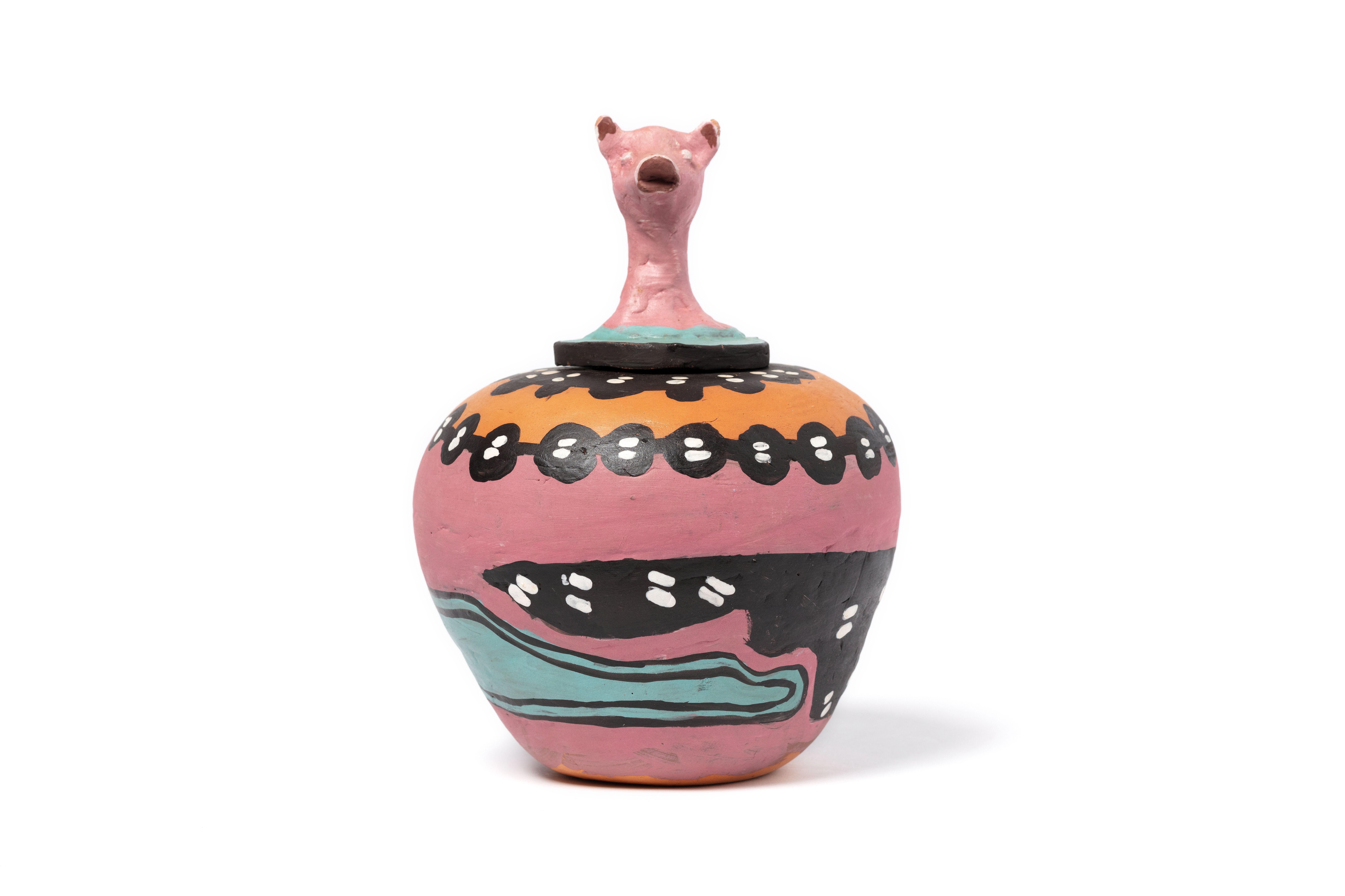 A round hand-formed pot in thick clay with a sculpted lid representing the head of a pig. Around the base is a painted pink ground, black and aqua forms, and white paint applied in the 'dot painting' style. The interior is glazed. Accompanying the pot is a folded, beige cardboard label. The label reads 'Hermannsburg Potters, 'My Pig Is Looking for Food', entirely hand crafted and painted by the Aranda people from Hermannsburg, Central Australia, Irene Entata.