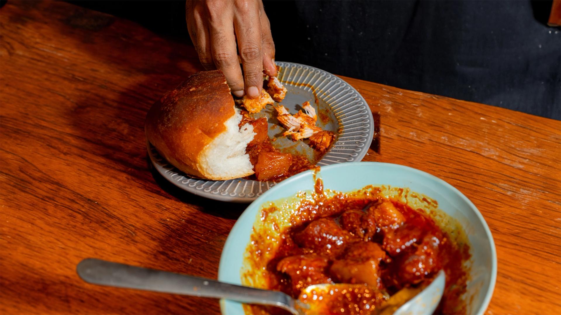 A brown hand mops up the last of an Indian curry with a half-eaten white bread roll.