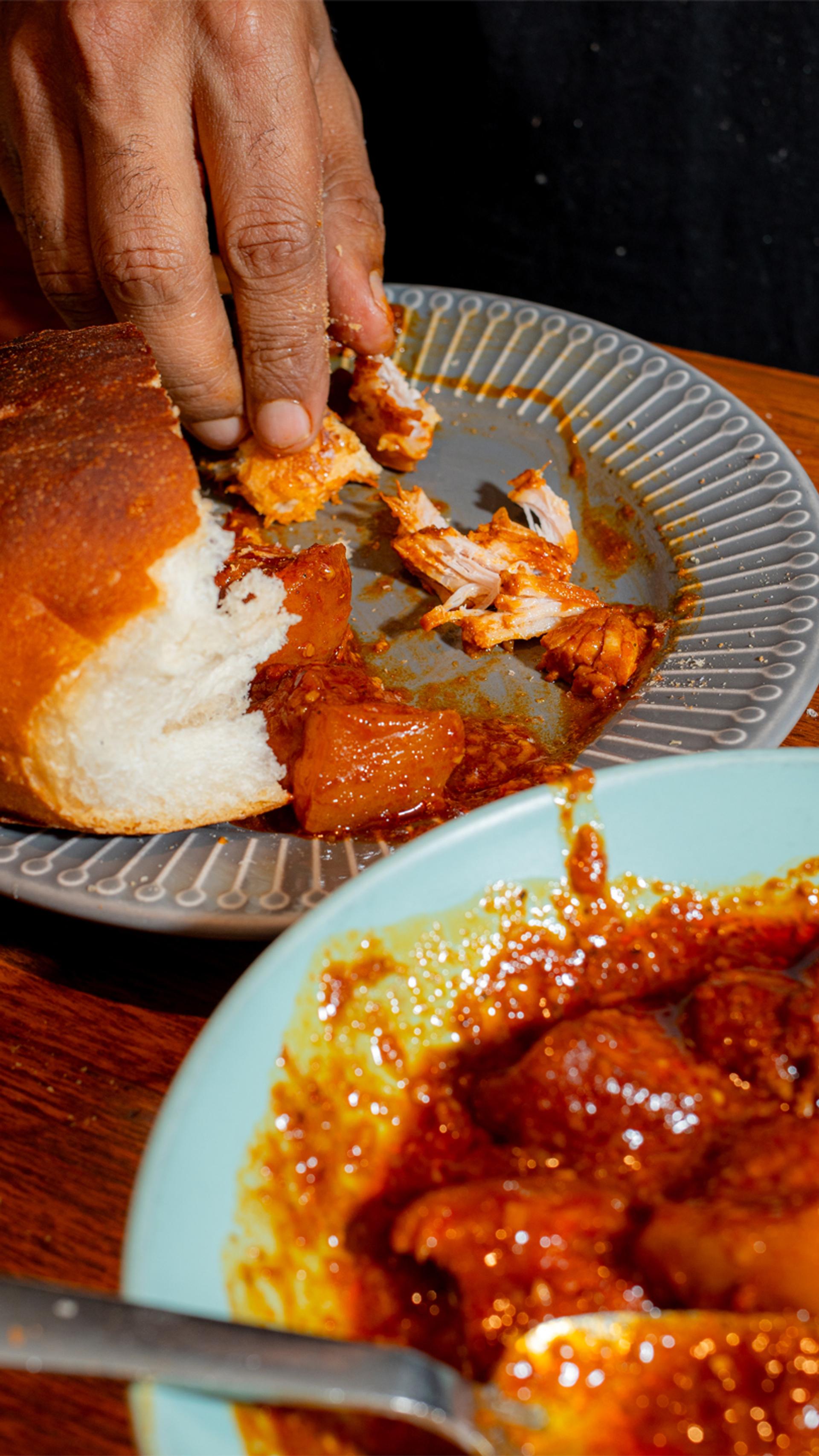 A brown hand mops up the last of an Indian curry with a half-eaten white bread roll.