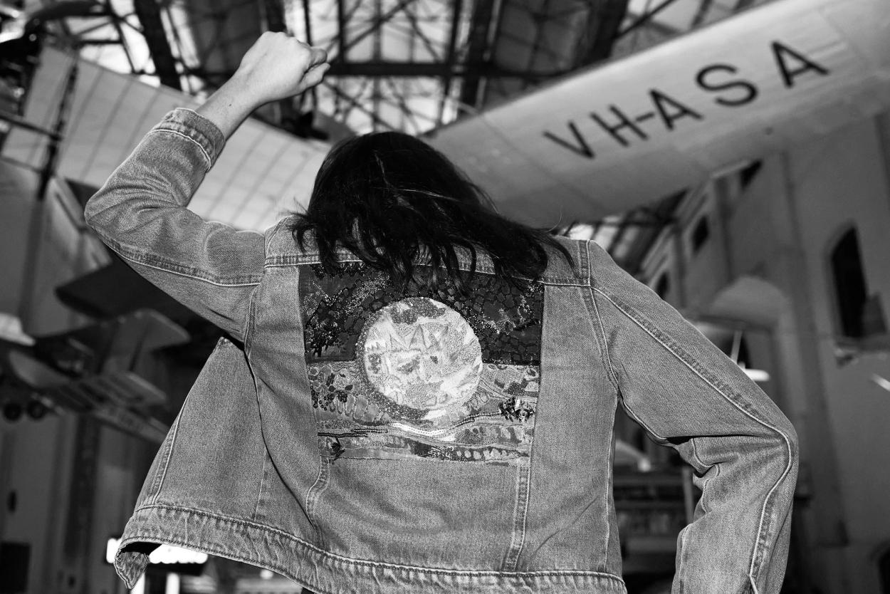 Black and white photograph. Mid shot of a person with their back to the camera, wearing a denim jacket with an applique of the Aboriginal flag. Their left arm and fist are raised in the air.