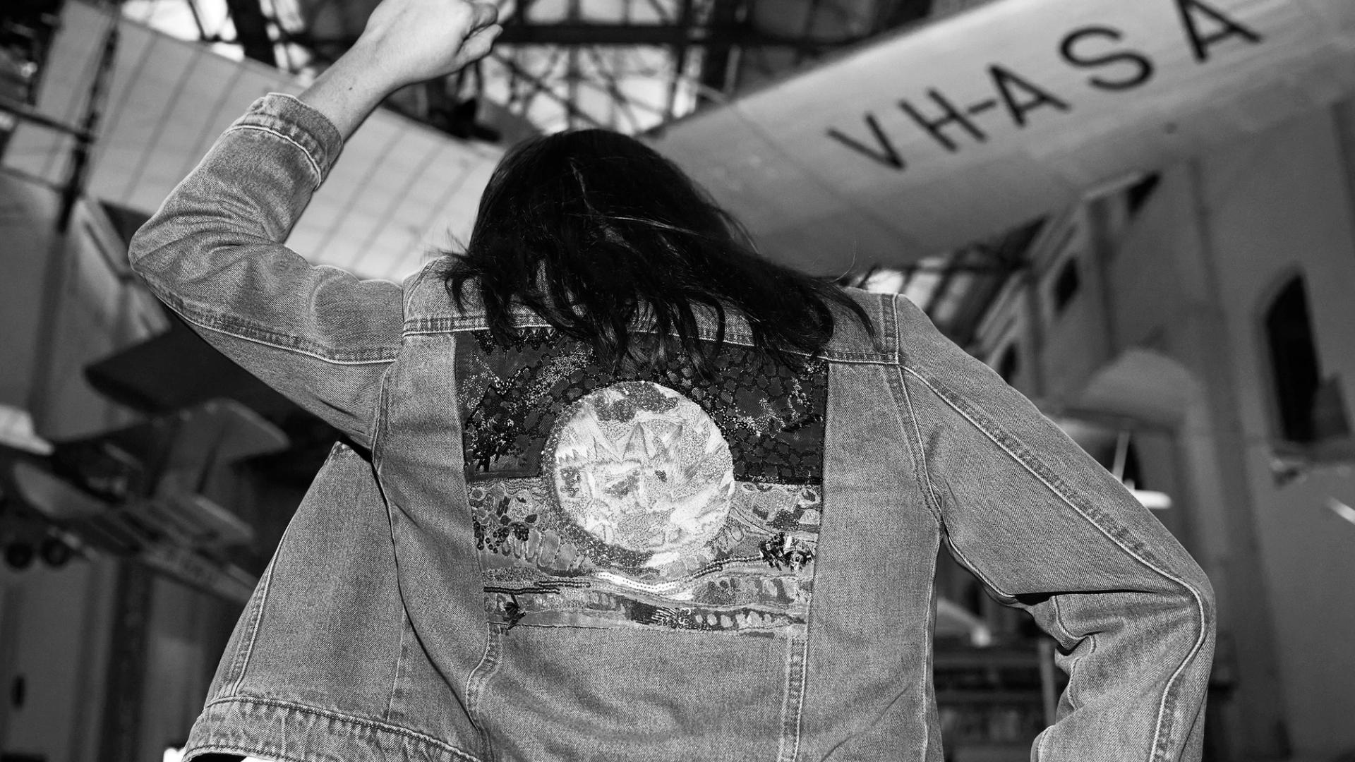 Black and white photograph. Mid shot of a person with their back to the camera, wearing a denim jacket with an applique of the Aboriginal flag. Their left arm and fist are raised in the air.
