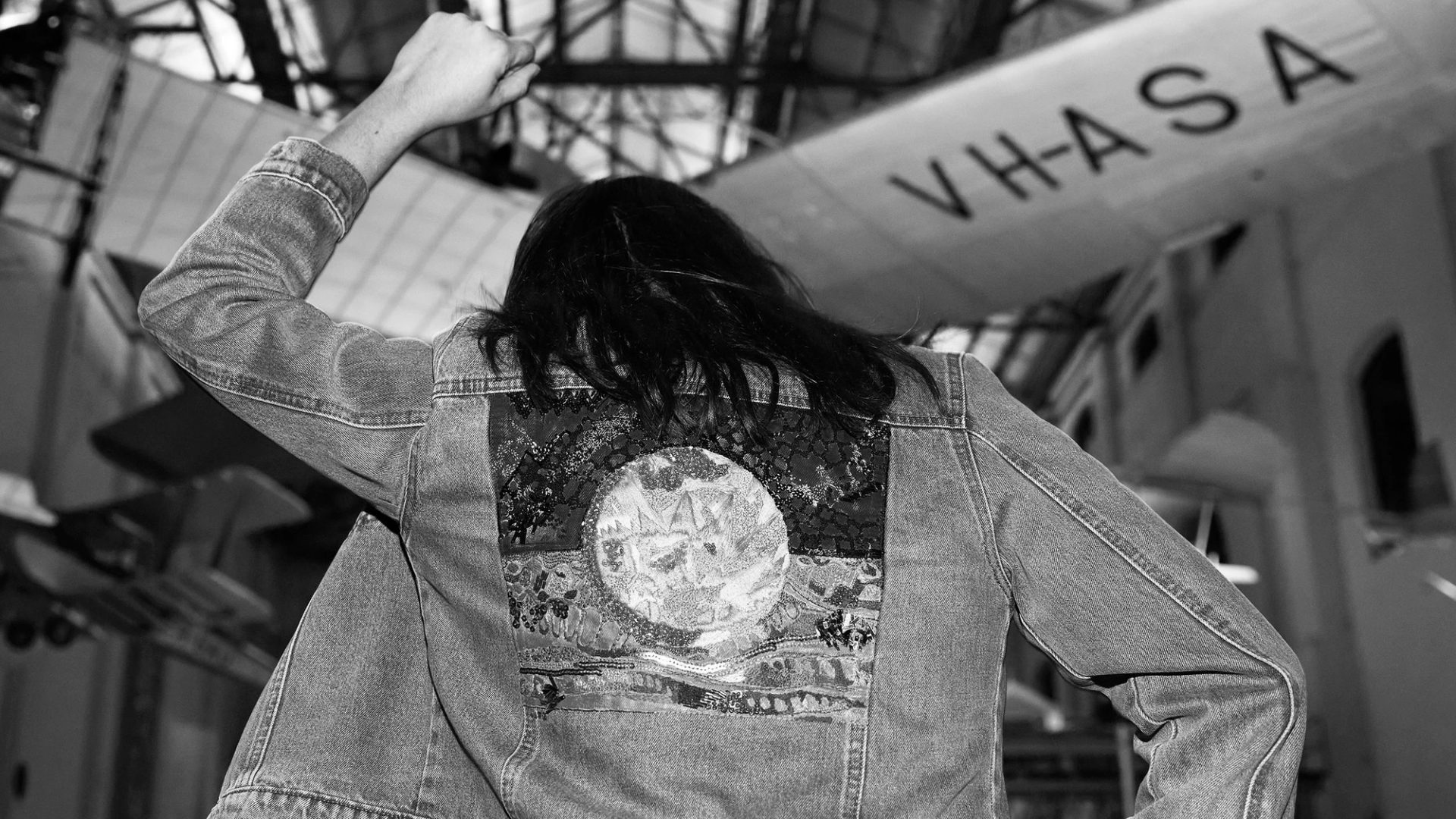 Black and white photograph. Mid shot of a person with their back to the camera, wearing a denim jacket with an applique of the Aboriginal flag. Their left arm and fist are raised in the air.