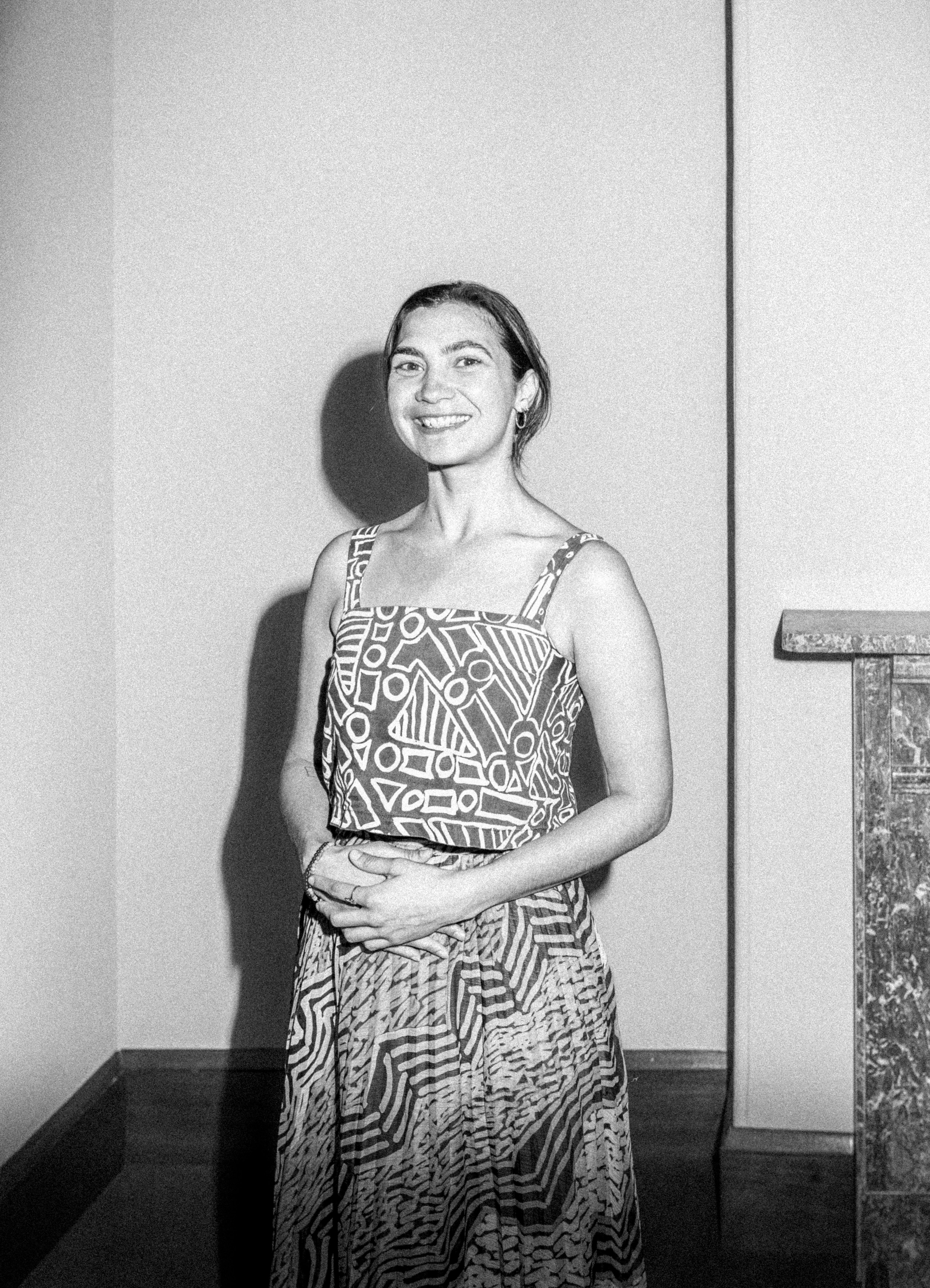 A woman with short hair wearing a strap sleeve shirt with intricate patterns (Rachael Hocking) stands against a white background, with the cameras flash casting a shadow. They are facing the camera with their hands are held together at her waist.