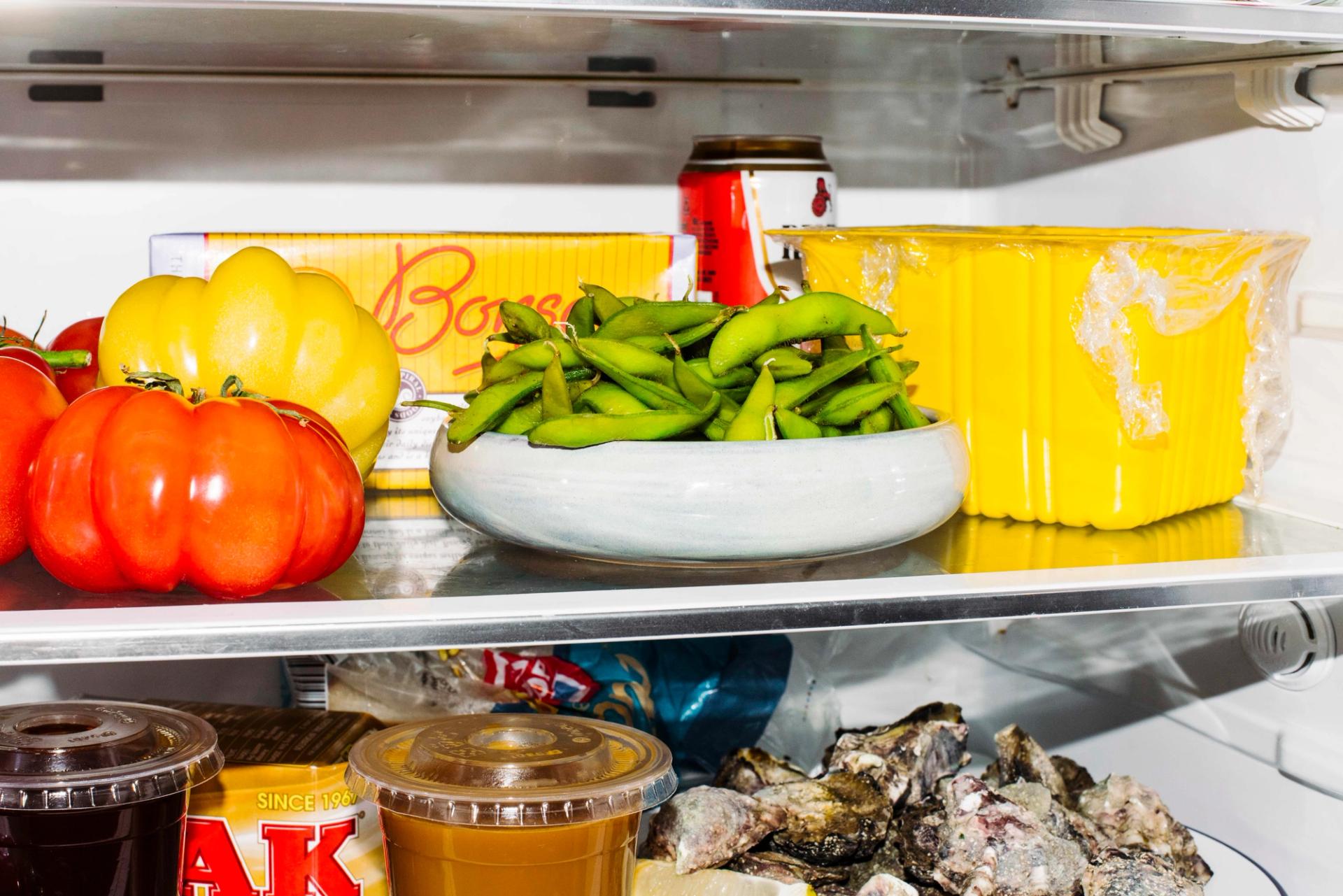 A small grey bowl filled with edamame beans sits on a middle fridge shelf. There is a red Resch’s beer can and a yellow Bonsoy carton behind it.