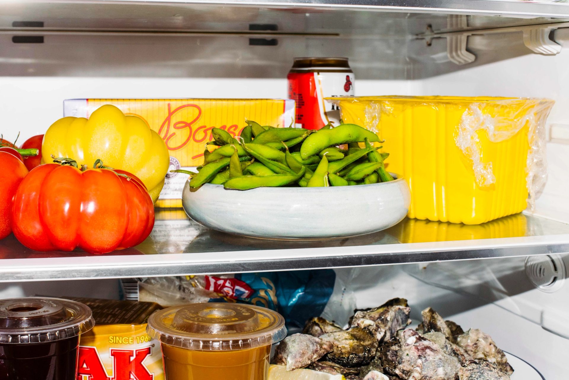 A small grey bowl filled with edamame beans sits on a middle fridge shelf. There is a red Resch’s beer can and a yellow Bonsoy carton behind it.