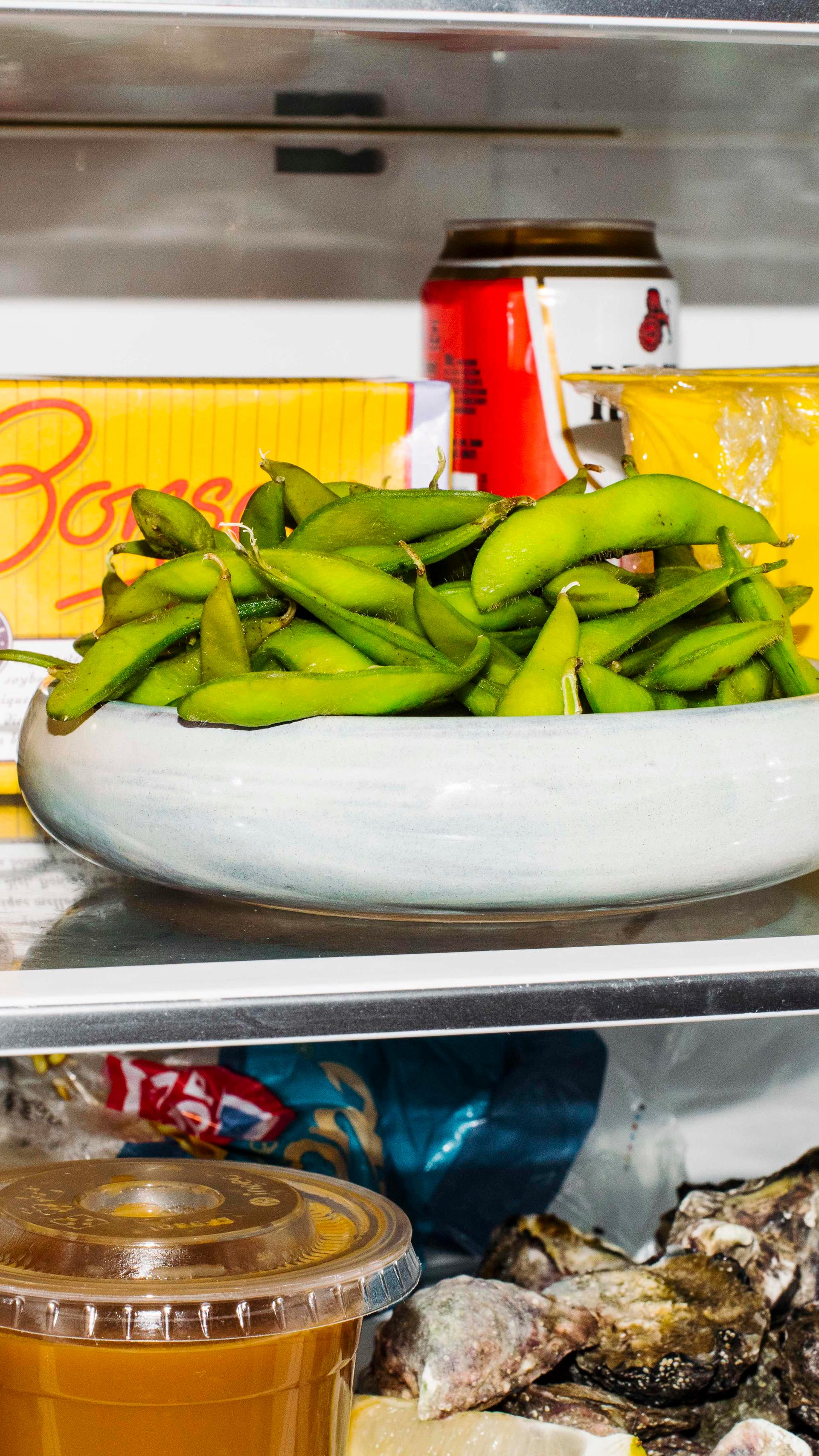 A small grey bowl filled with edamame beans sits on a middle fridge shelf. There is a red Resch’s beer can and a yellow Bonsoy carton behind it.