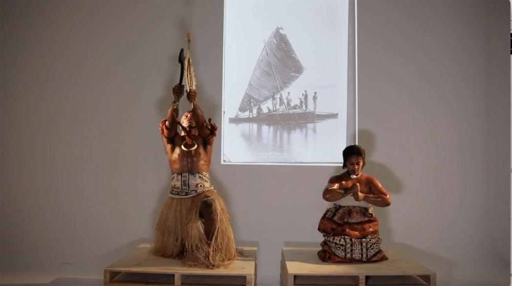 Two people wearing traditional Fijian dress are mid performance in front of a projected image of a historical Fijian canoe (waqa).