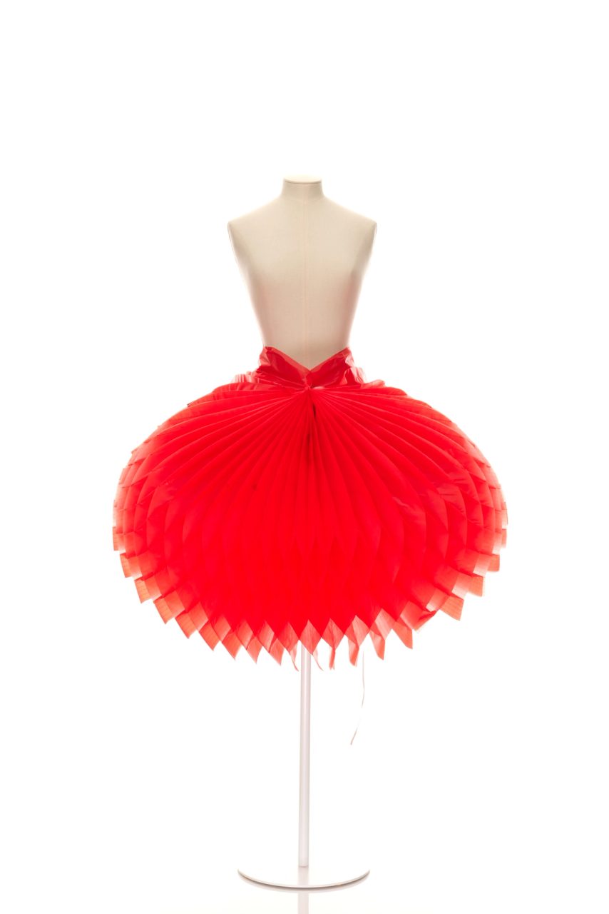 Skirt and slip cover from 'The Wizard of Jeanz' collection, consists of a red skirt with press studs and ruffles stitched to the inside spine of a denim-look fabric book. The front of the book has a silver border and bears the text 'The Wizard of Jeanz'. This text is also printed on the spine of the book, in addition to the surname of the designer 'Ohya' and a number 20' which represents the volume number in the series. A black fabric label with the text 'Ohya' is sewn into the waistband of the skirt. A grey cord book mark is also sewn into the spine of the book.The garments are folded to fit into a silver cardboard slip cover which is embossed along the spine with the text 'The Wizard of Jeanz' and the surname of the designer.