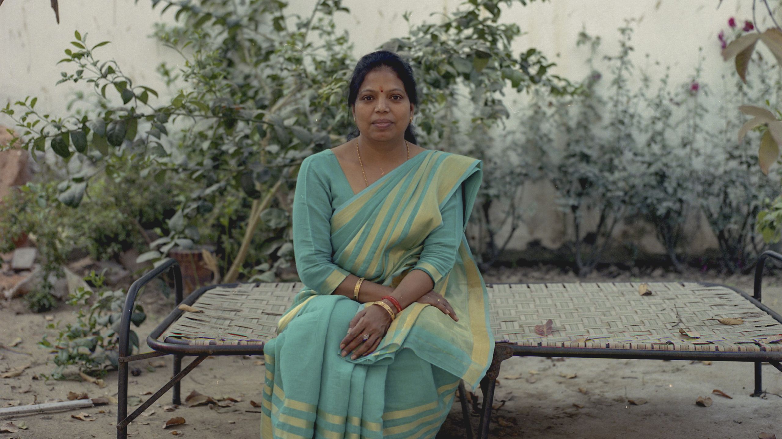 A person wearing a blue and yellow/gold sari sitting on a metal bed frame outside, behind them is a beige wall with green shrubbery along the bottom.