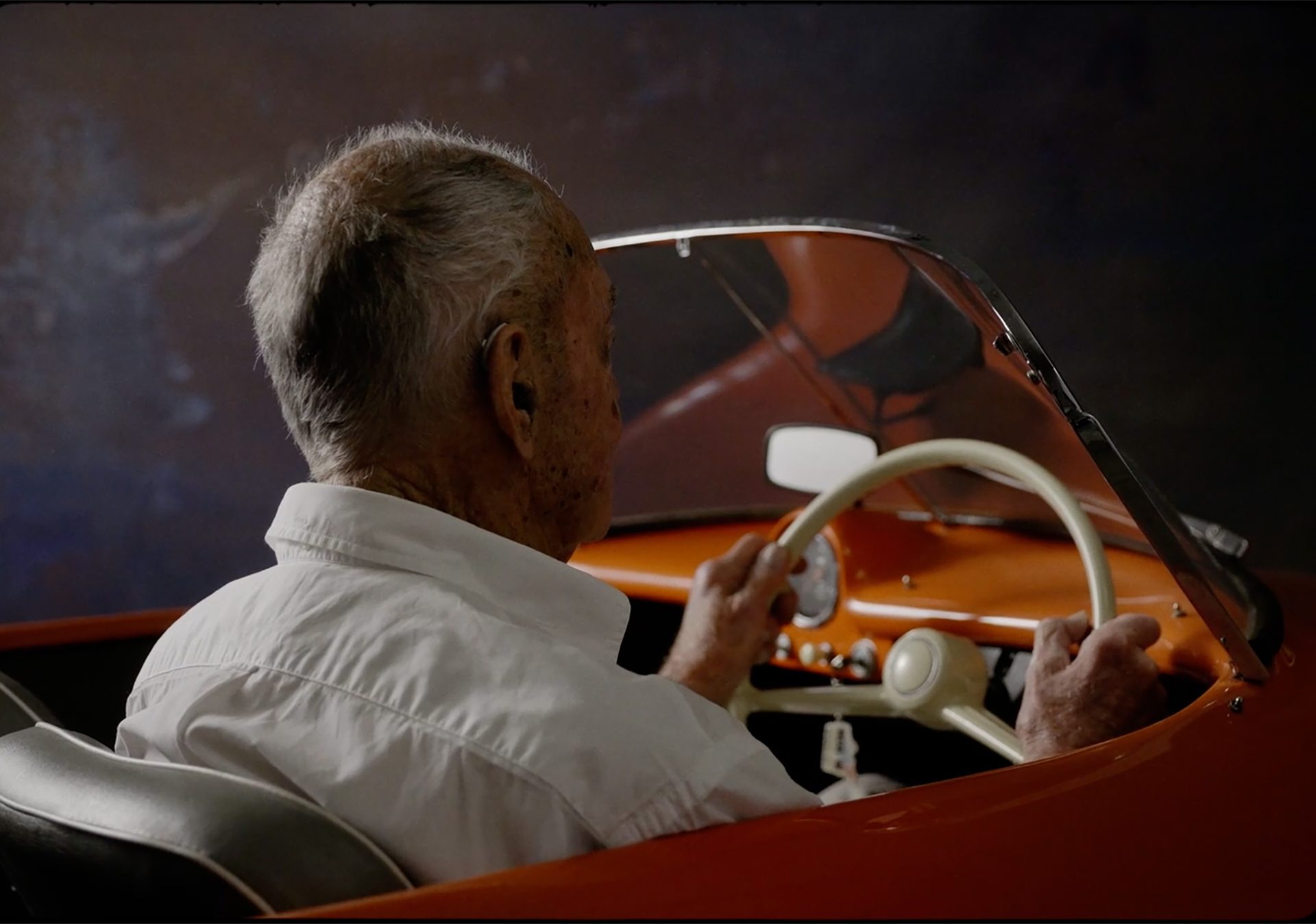 Figure sits behind the steering wheel of an orange open roof car
