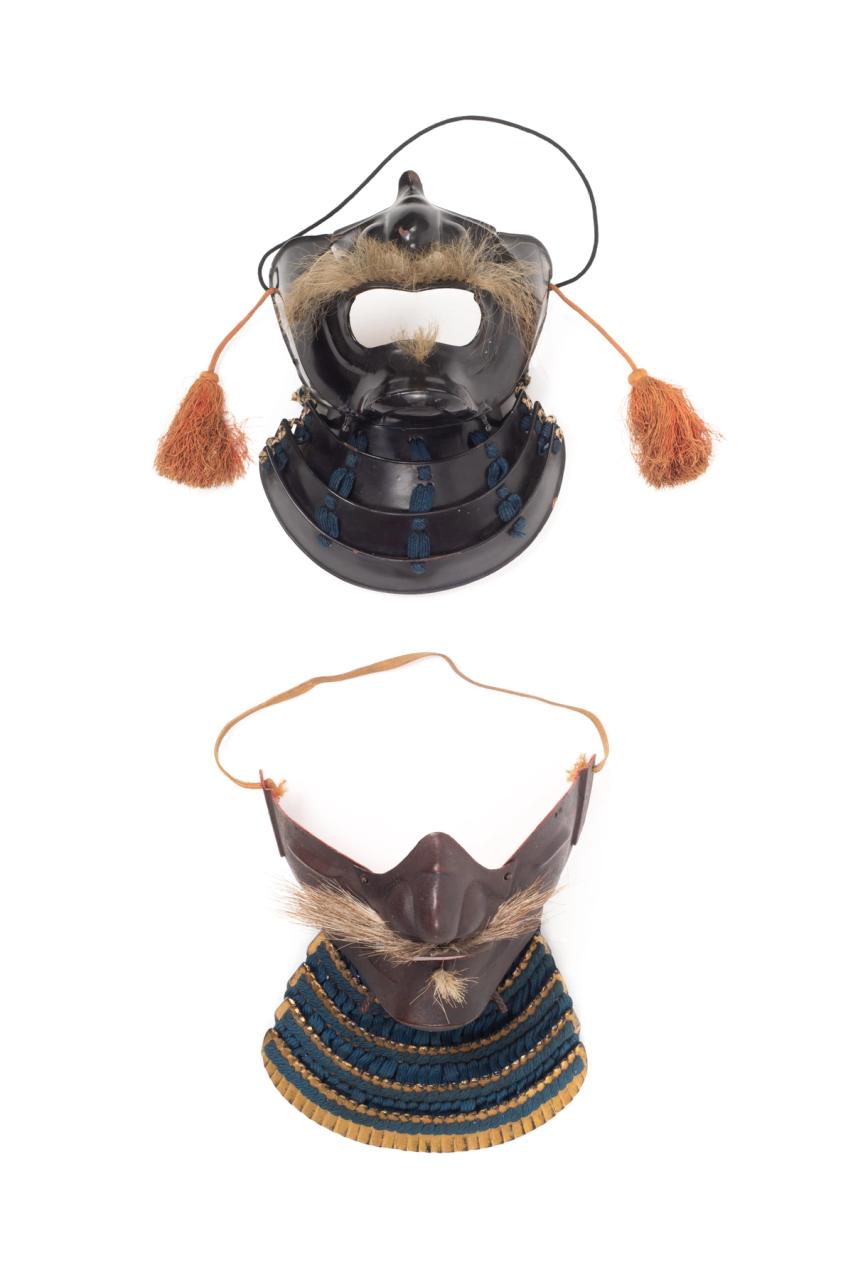 Two masks as Japanese facial armour (Mempō). One mask features a blue fabric cordage neck guard, or Yodare-kake, chin, cheek and nose armour and a horse hair moustache and small beard tuft. The second mask features a highly lacquered leather Yodare-kake with blue cordage and highly lacquered chin, cheek and nose armour, with a large mouth opening, a horse hair moustache and beard tuft and tassels, or Agemaki attached to each cheek guard.