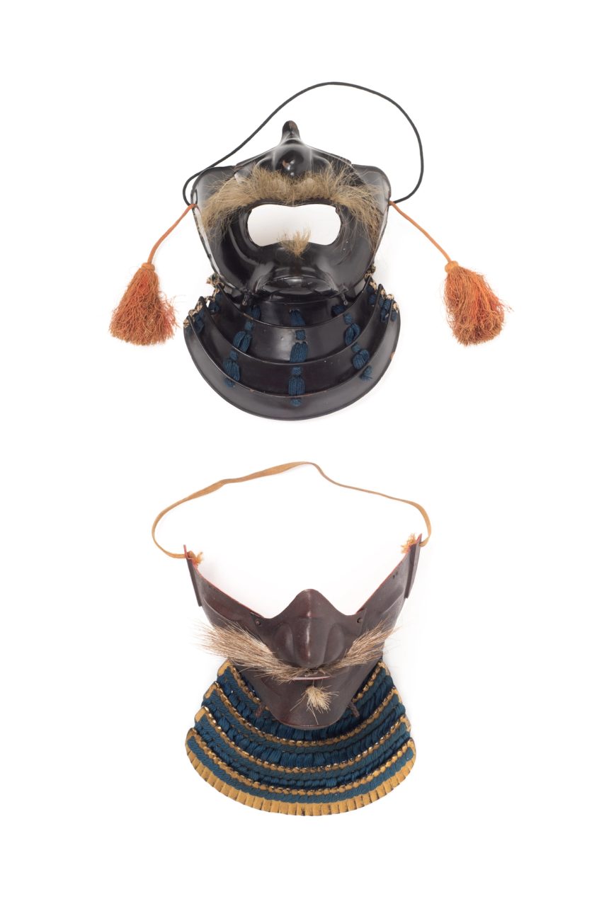 Two masks as Japanese facial armour (Mempō). One mask features a blue fabric cordage neck guard, or Yodare-kake, chin, cheek and nose armour and a horse hair moustache and small beard tuft. The second mask features a highly lacquered leather Yodare-kake with blue cordage and highly lacquered chin, cheek and nose armour, with a large mouth opening, a horse hair moustache and beard tuft and tassels, or Agemaki attached to each cheek guard.