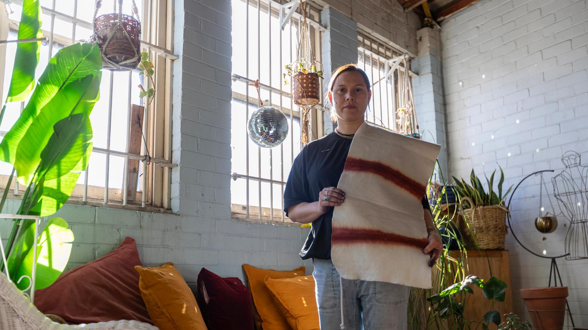 A woman holding a piece of textile, in a brick wall room, with windows behind.