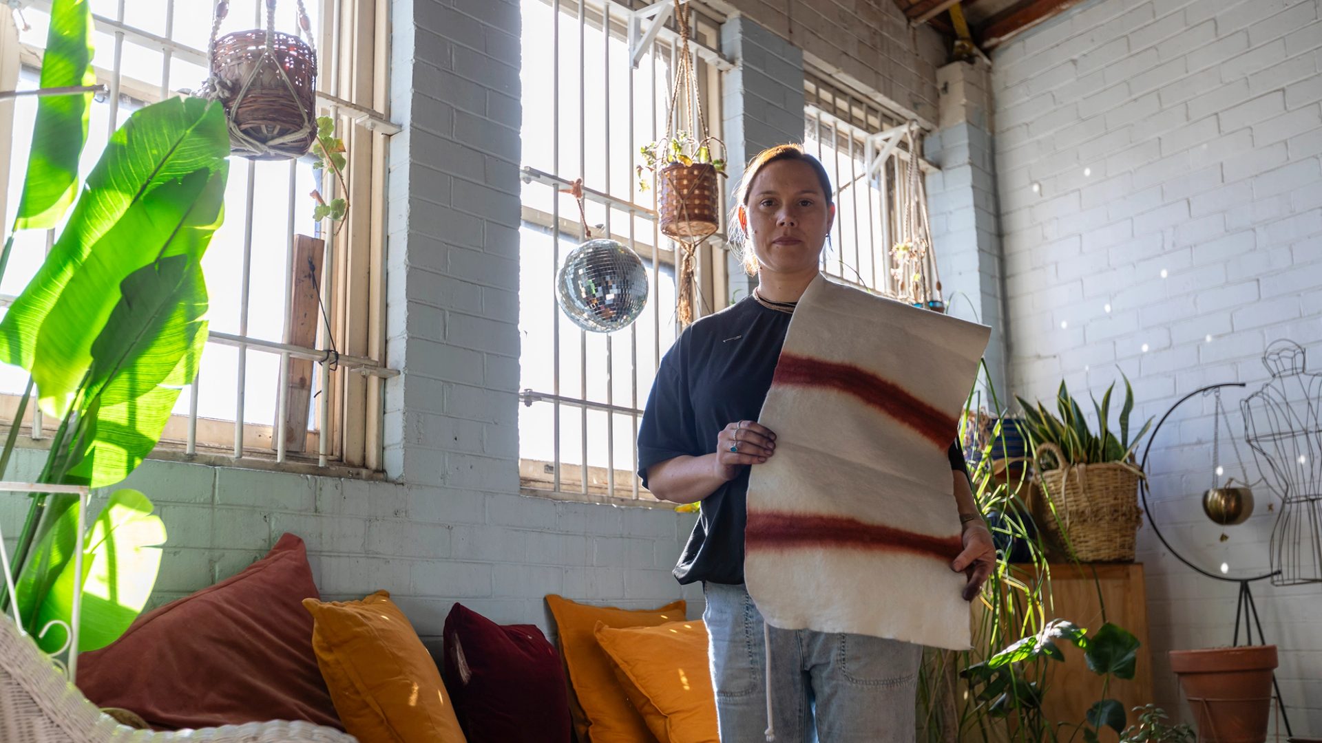 A woman holding a piece of textile, in a brick wall room, with windows behind.