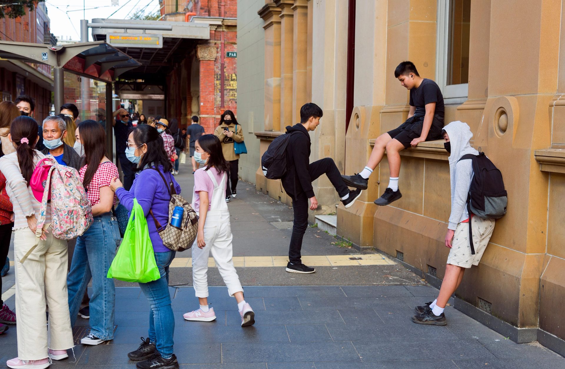 A crowd of people at the Haymarket tram stop, a kid sits on the window ledge of the sandstone building next to the tram stop.