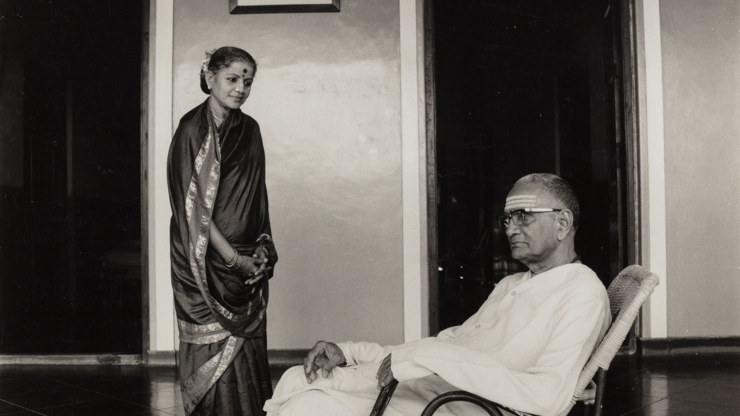 Black and white photograph depicting a woman (Subbulakshmi) wearing a sari looking down at a man (her husband T. Sadasivam) seated.