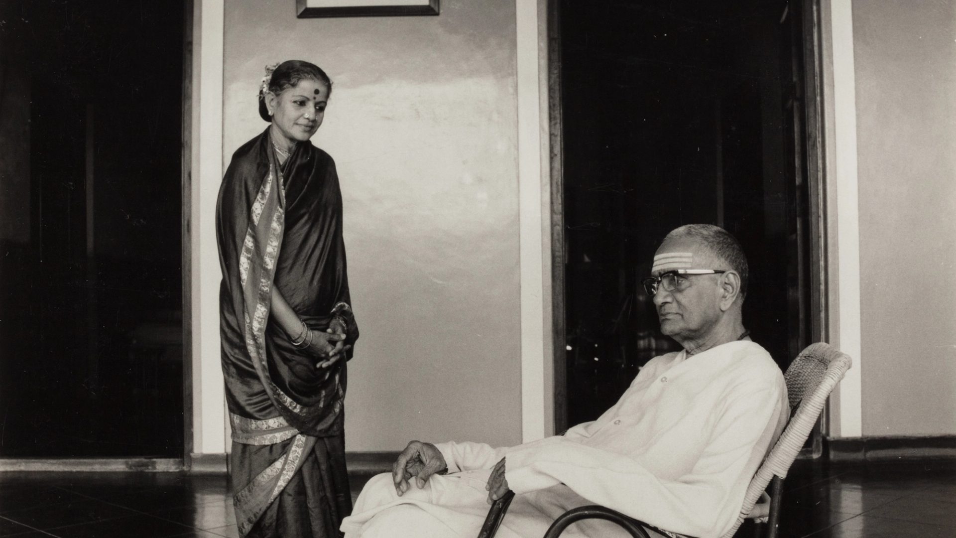 Black and white photograph depicting a woman (Subbulakshmi) wearing a sari looking down at a man (her husband T. Sadasivam) seated.