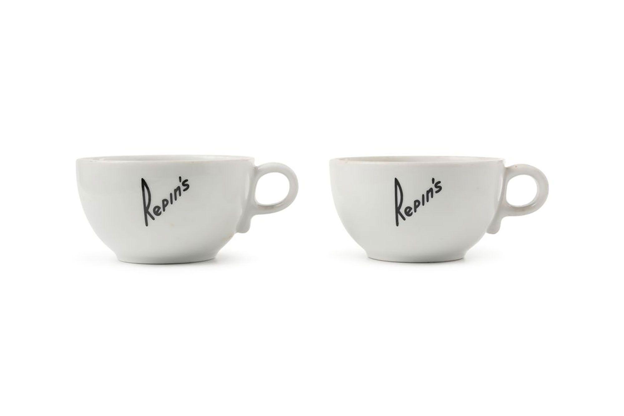 Two white porcelain coffee cups. The cups have a short base, a rounded bowl and a small circular handle. 'REPIN'S' is printed in black writing diagonally across the sides of the cups. Marked on base.