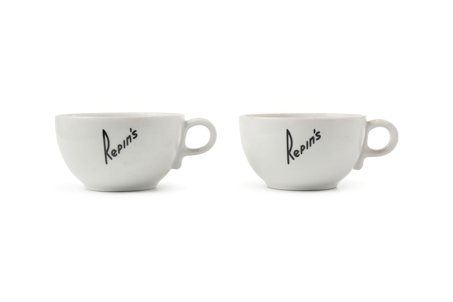 Two white porcelain coffee cups. The cups have a short base, a rounded bowl and a small circular handle. 'REPIN'S' is printed in black writing diagonally across the sides of the cups. Marked on base.