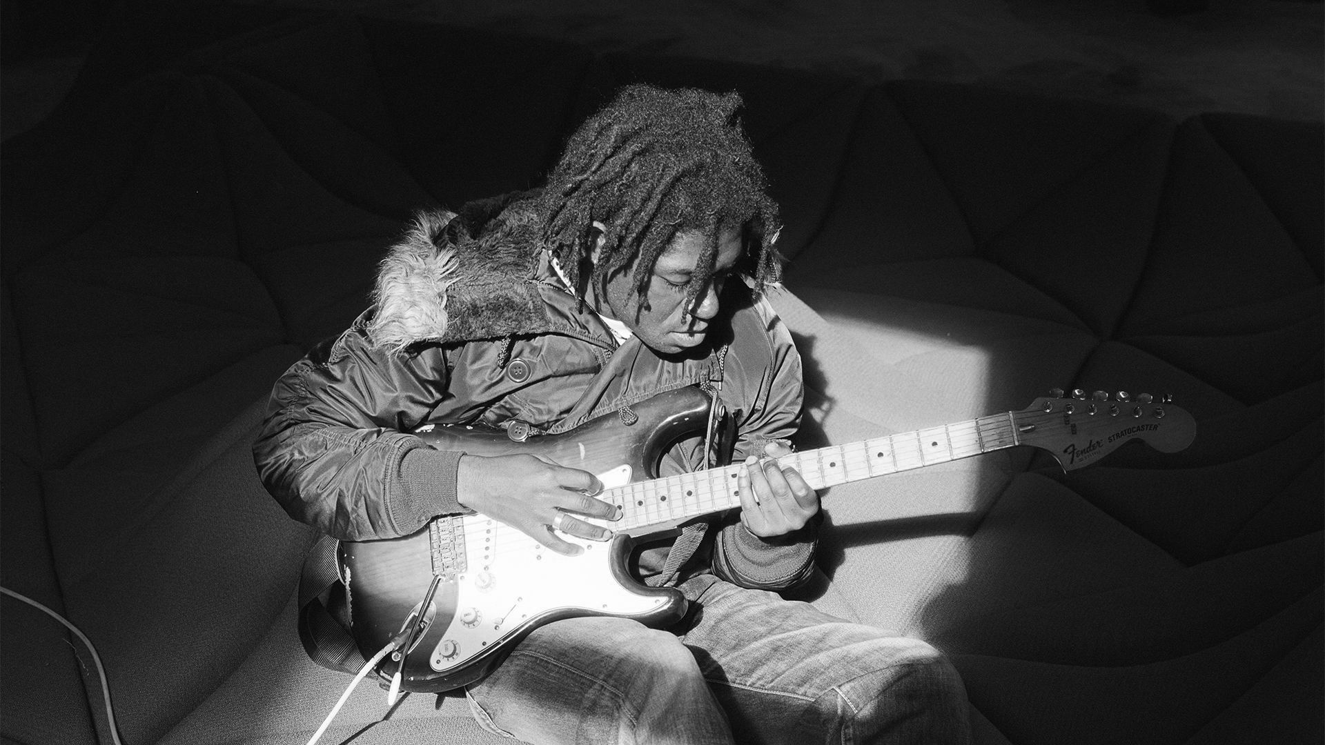 The musician Friday*, playing the guitar on a couch, photographed by Christine Yau