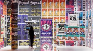 A large wall covered in a variety of brightly coloured band posters. A woman dressed in black is looking at the wall.